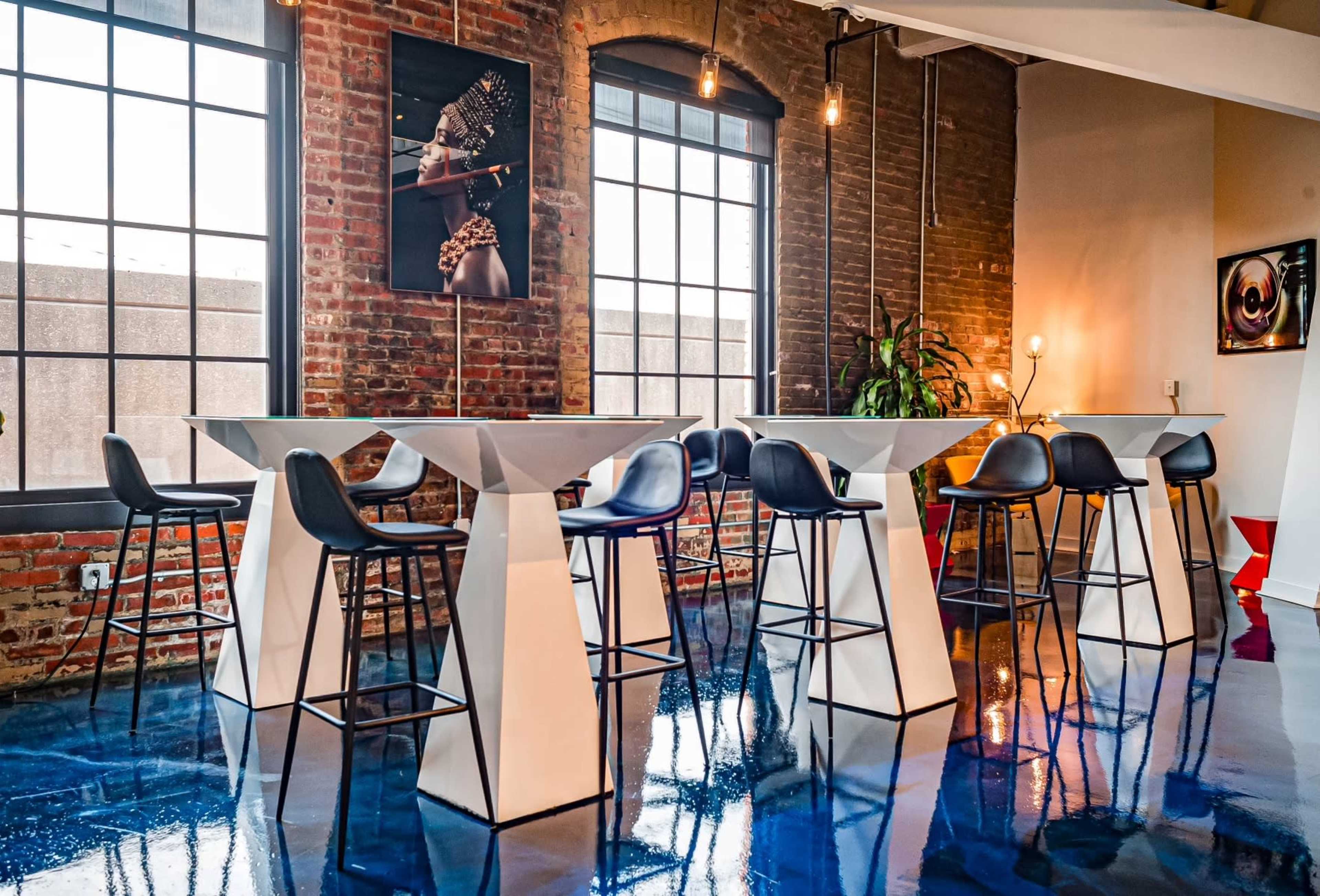 A modern interior with several white high tables and black bar stools against a backdrop of exposed brick walls and large windows.