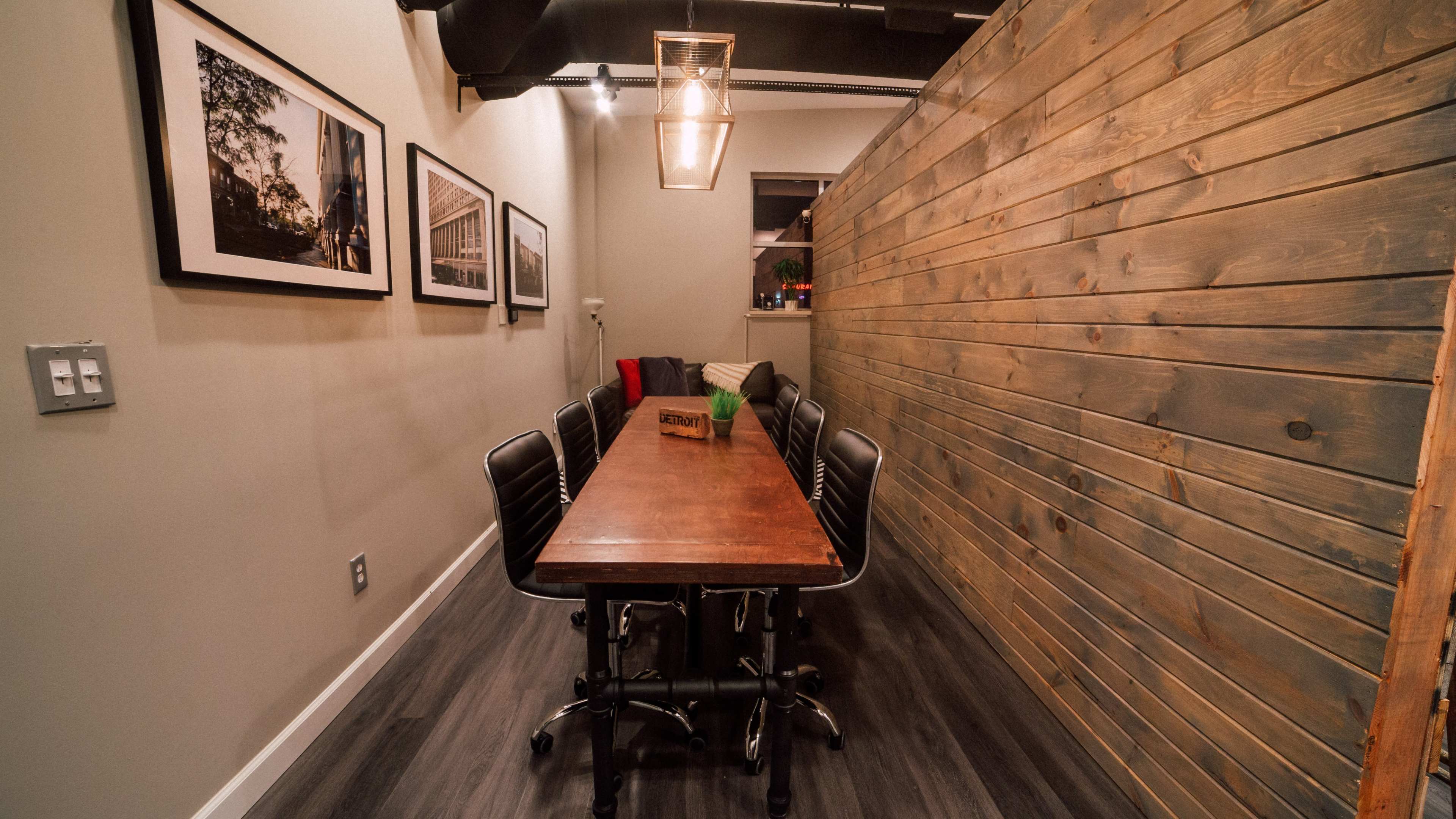 A long wooden table with black chairs is positioned in a narrow room featuring a wall made of wood planks and framed photographs on the adjacent wall.