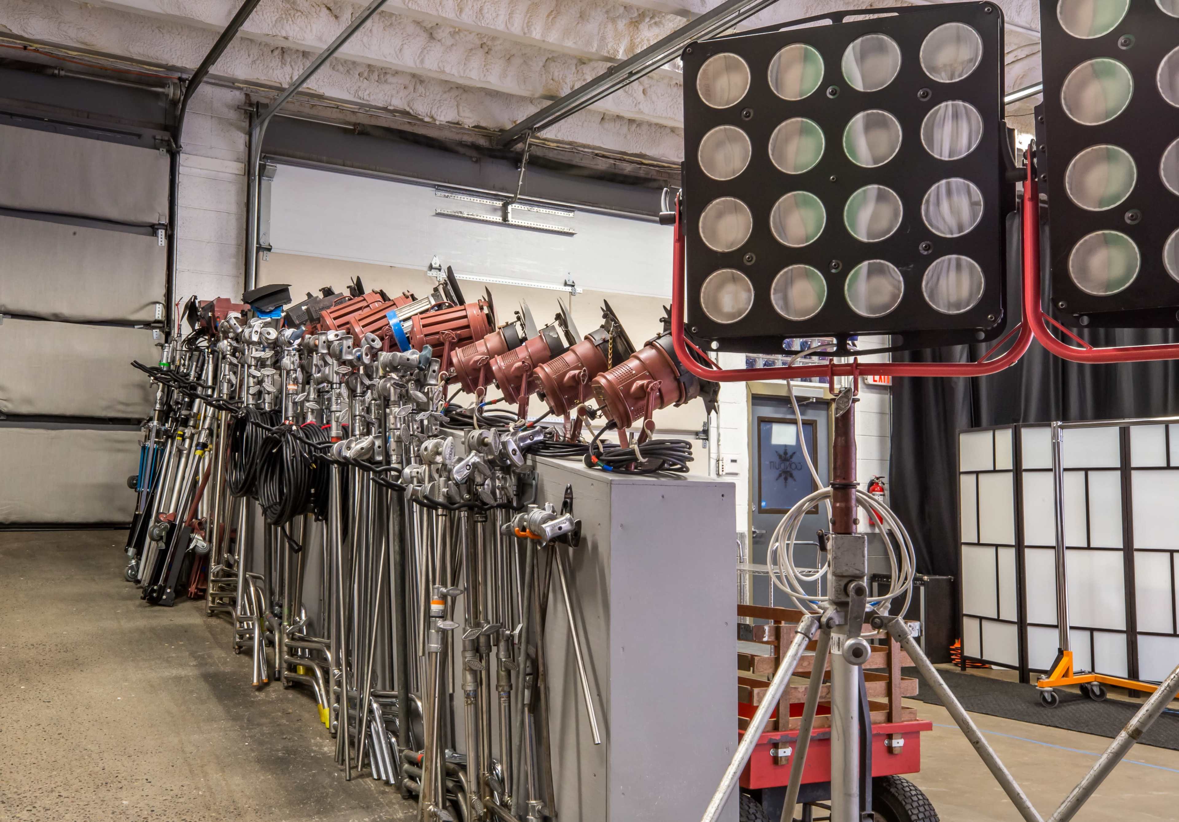 A row of lighting equipment, including multiple lamps and stands, is organized against the wall of a garage or warehouse.