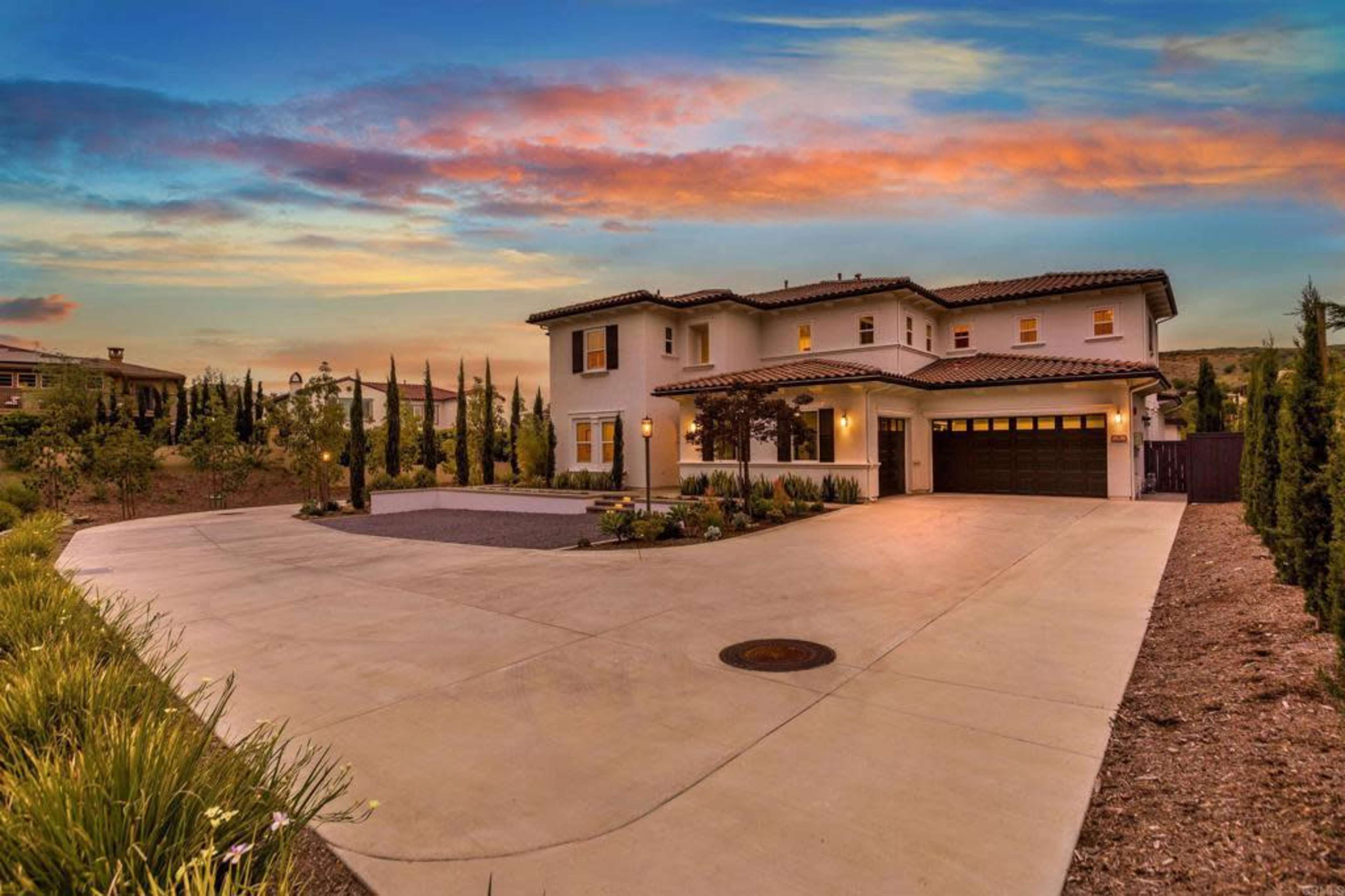 A two-story house with a tile roof and a landscaped front yard is set against a sunset sky.