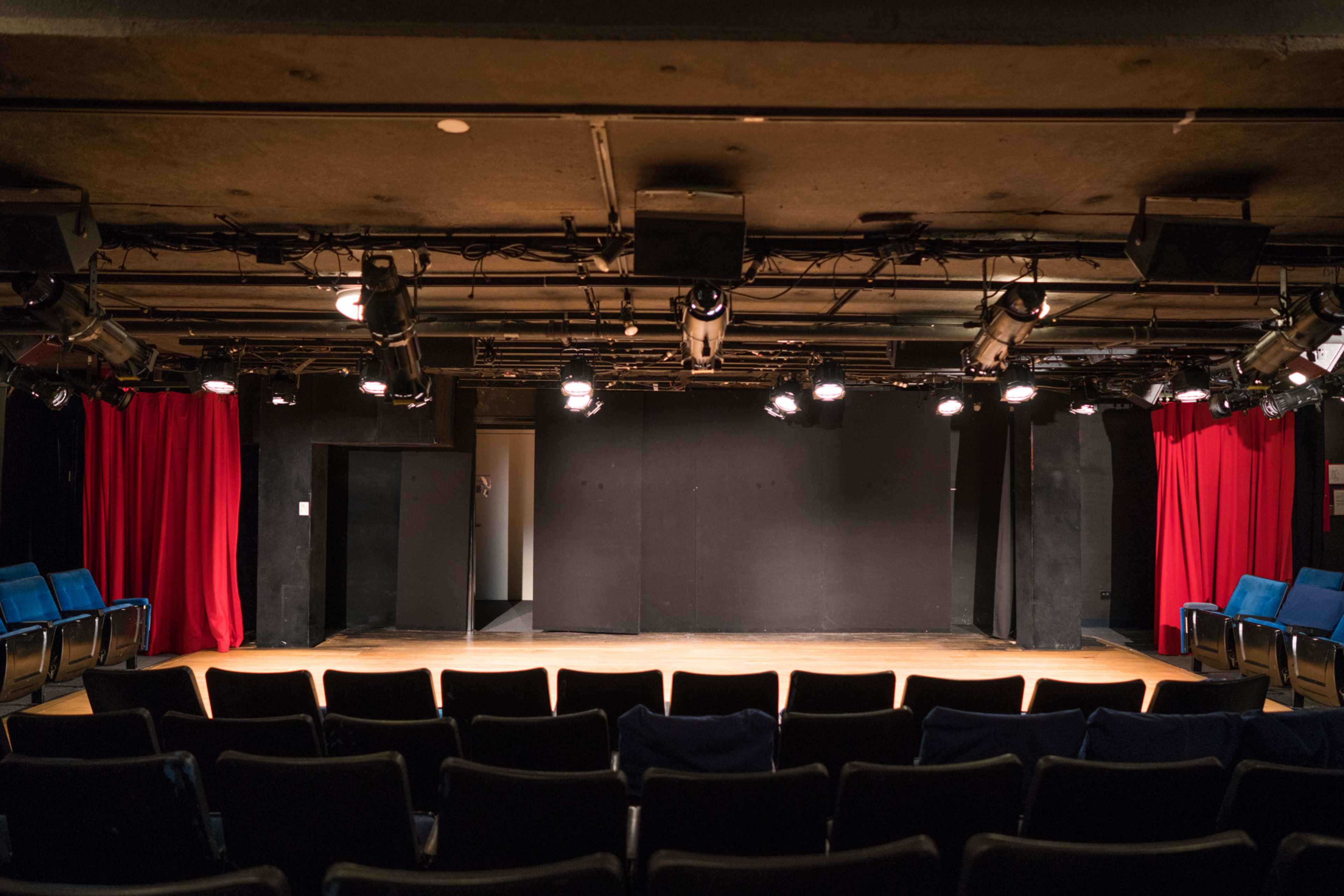 The image shows an empty theater stage with red curtains and rows of black seats facing the performance area.