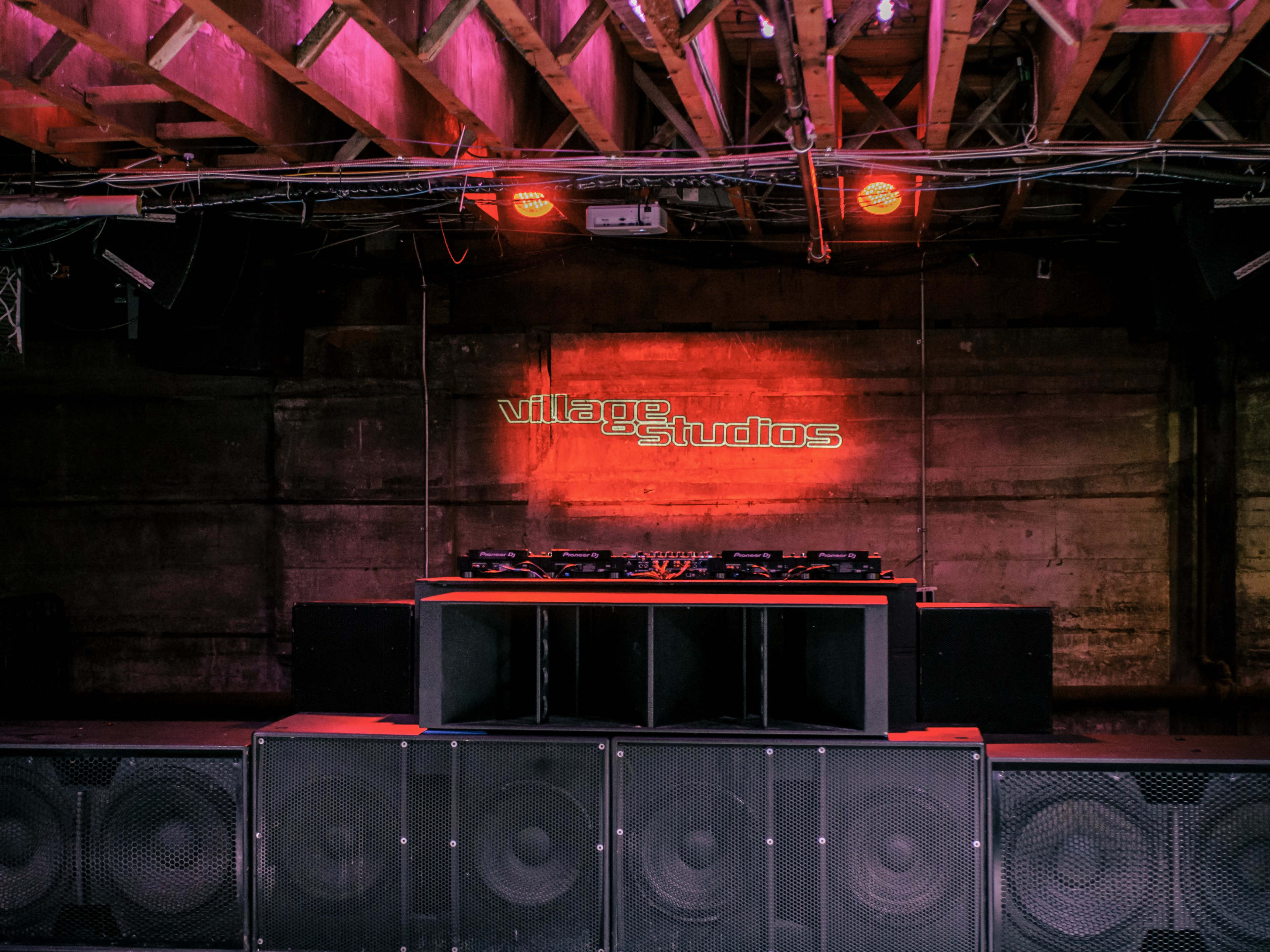 A DJ setup with turntables and speakers in a venue named "Village Studios," illuminated by pink lighting and a backlit sign.