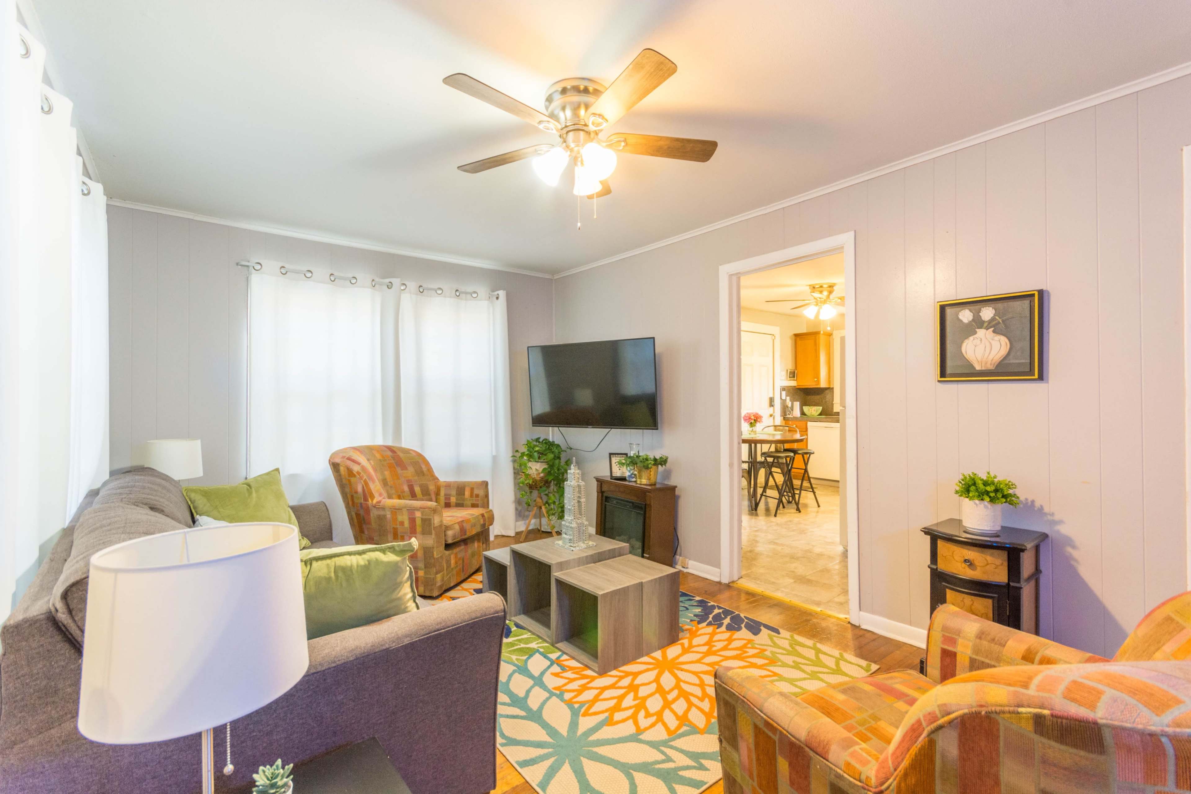 The living room features a gray sofa, two patterned chairs, a ceiling fan, a television mounted on the wall, and a coffee table, all set against light-colored walls and a colorful area rug.