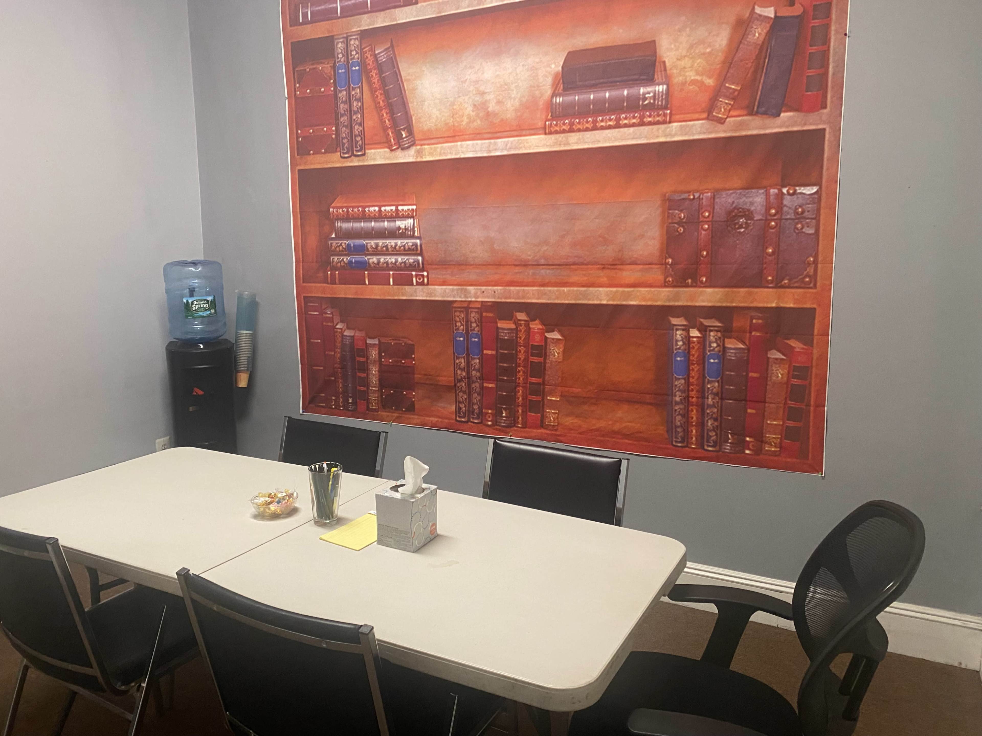 A conference room features a table with chairs, a water dispenser, and a wall mural of bookshelves lined with books.