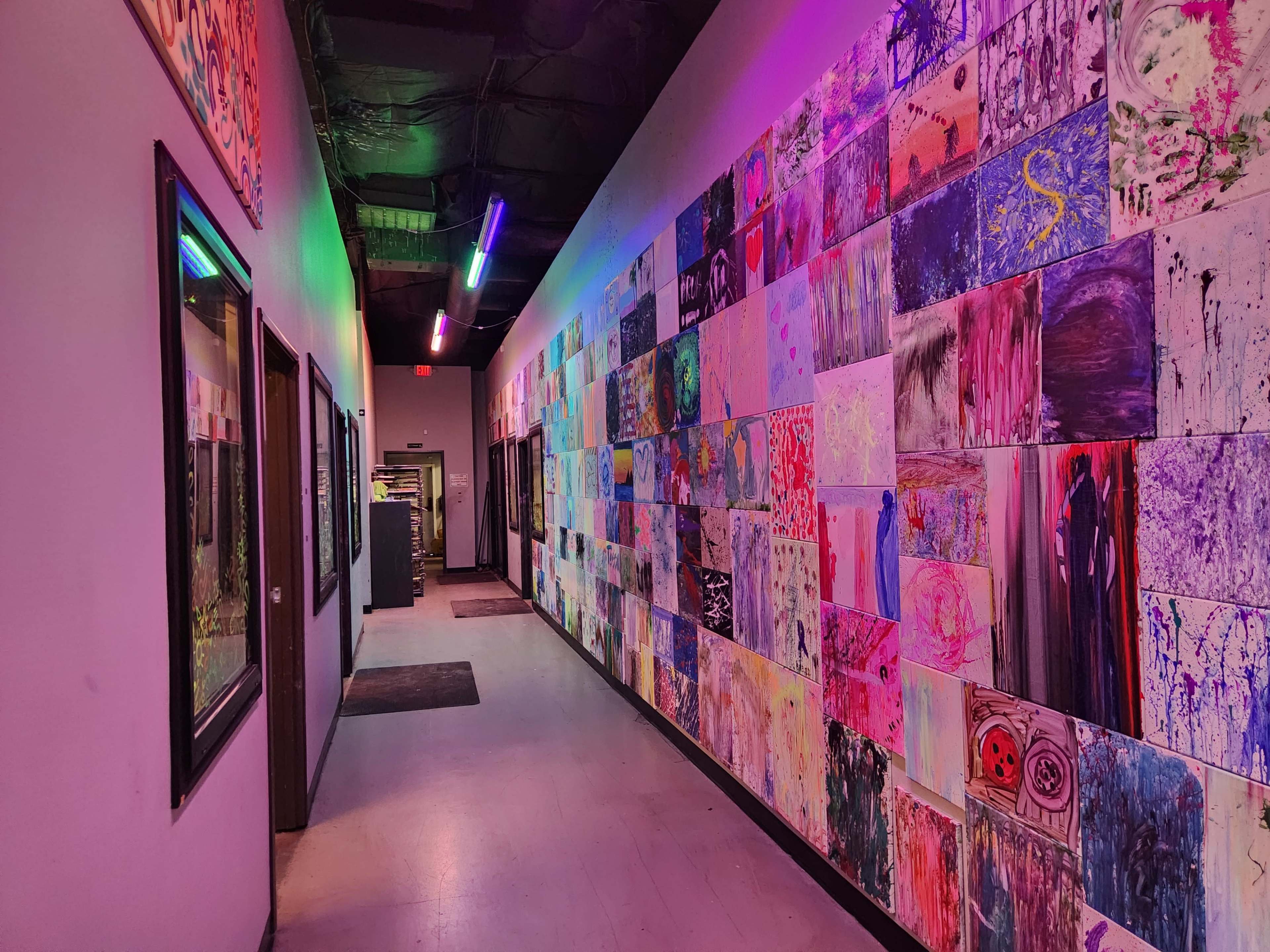 The image depicts a colorful hallway adorned with a variety of art pieces on one wall and illuminated by vibrant neon lights.