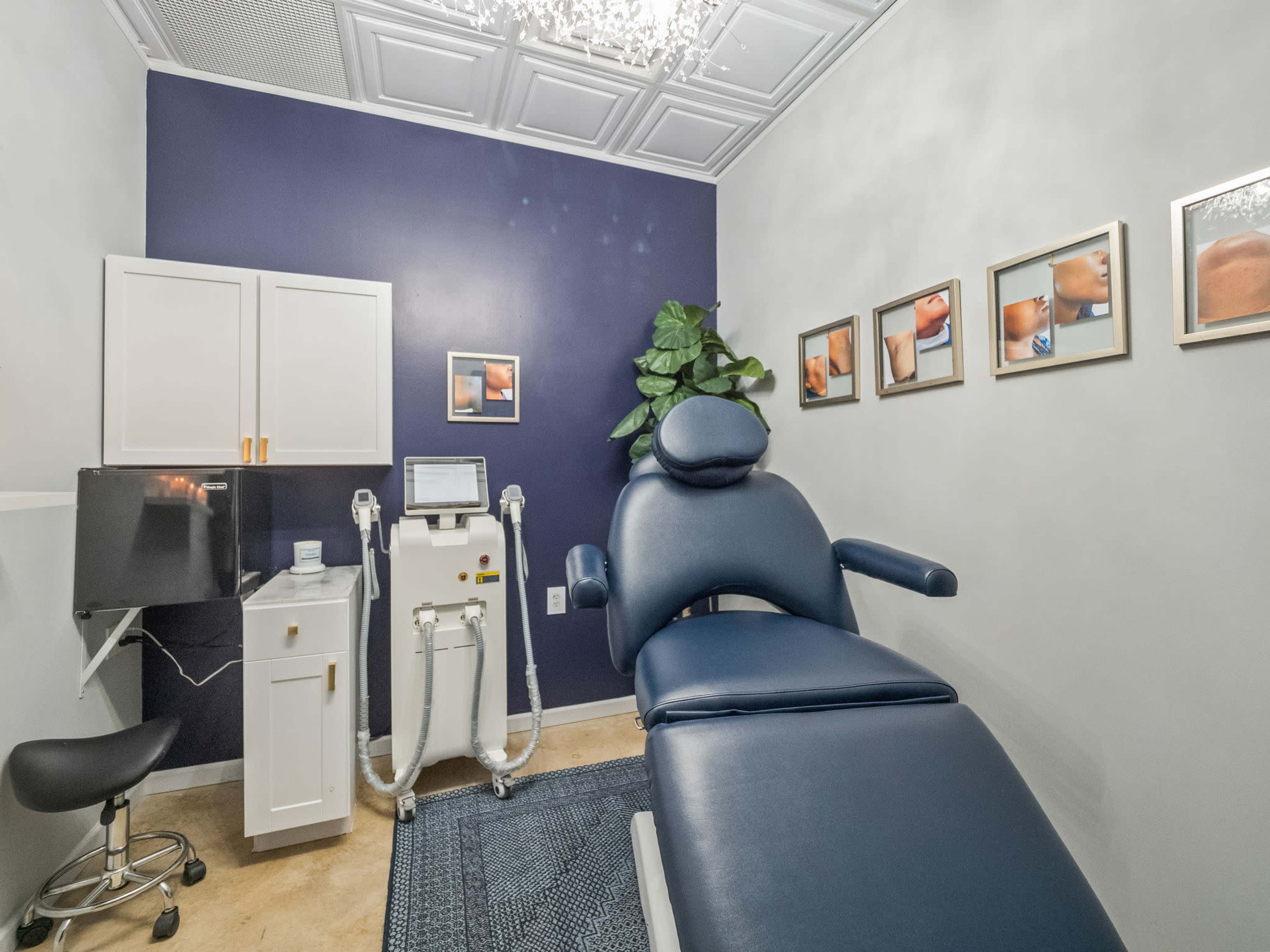 A treatment room features a medical chair, a monitor, and a machine against a dark blue wall adorned with framed photos.