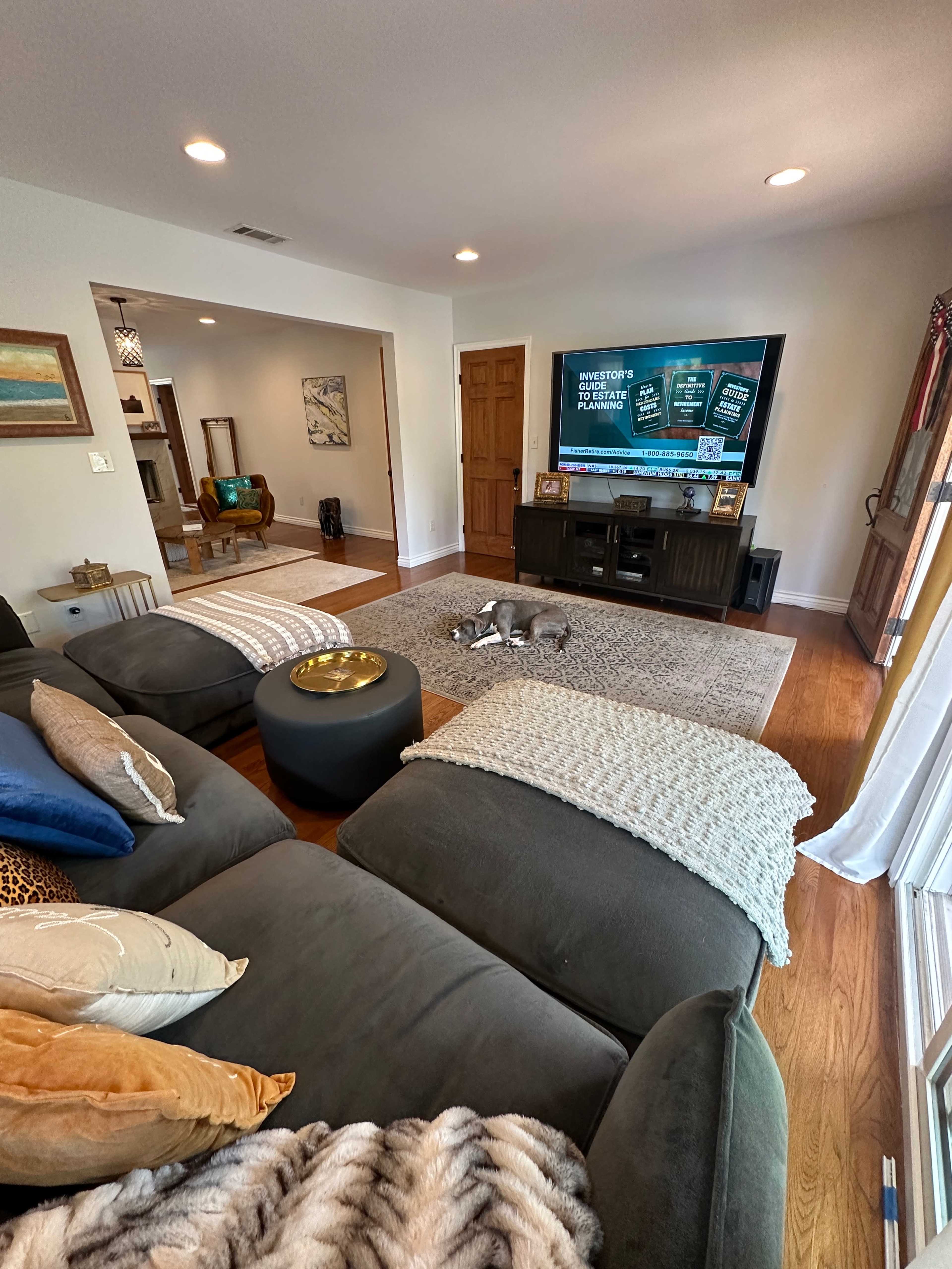 A spacious living room features a large sectional sofa, a coffee table, and a television displaying information, with hardwood floors and light-colored walls.