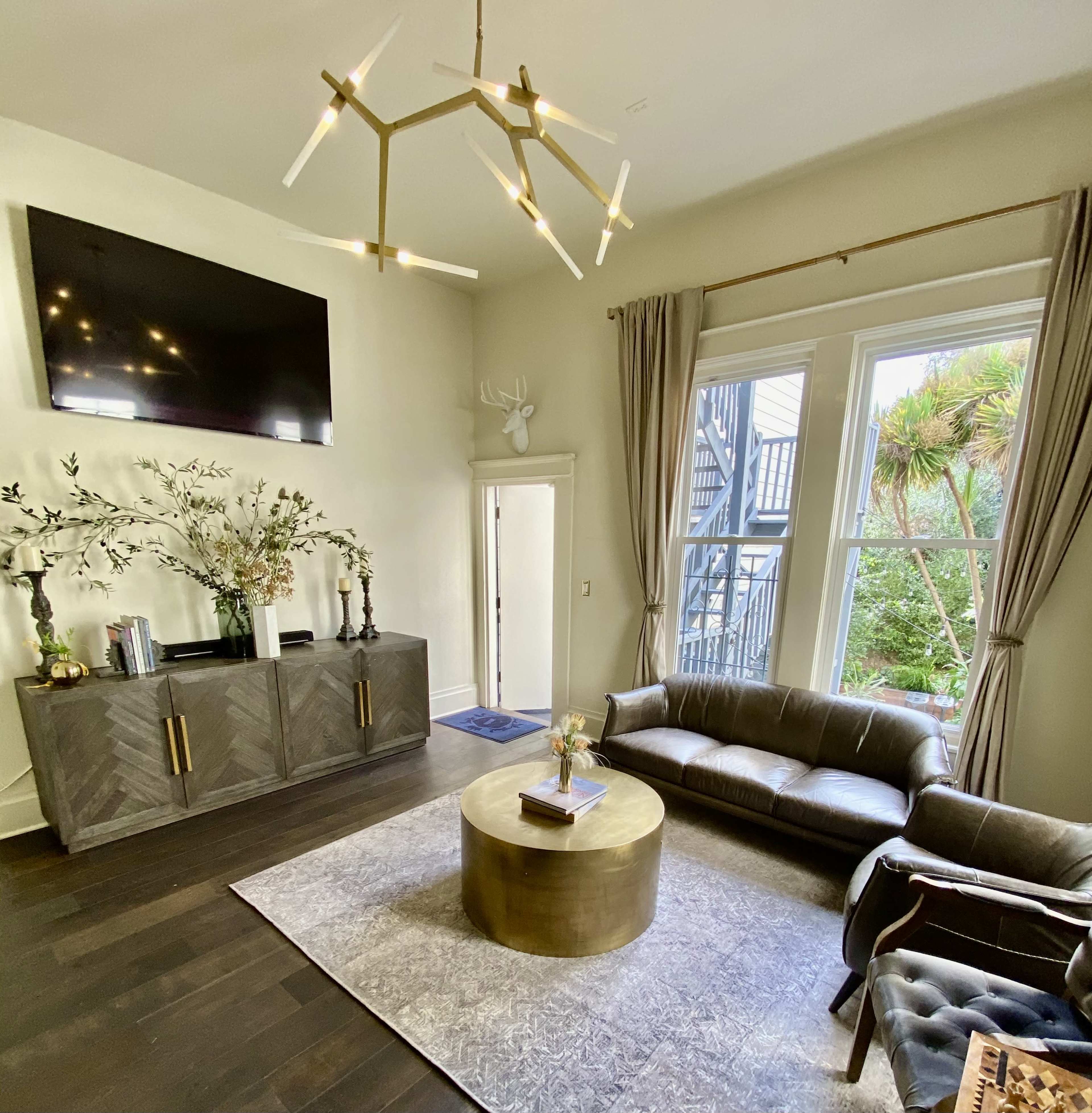 A modern living room features a television above a dark cabinet, a brown leather sofa, a round gold coffee table, and large windows with a view of greenery.