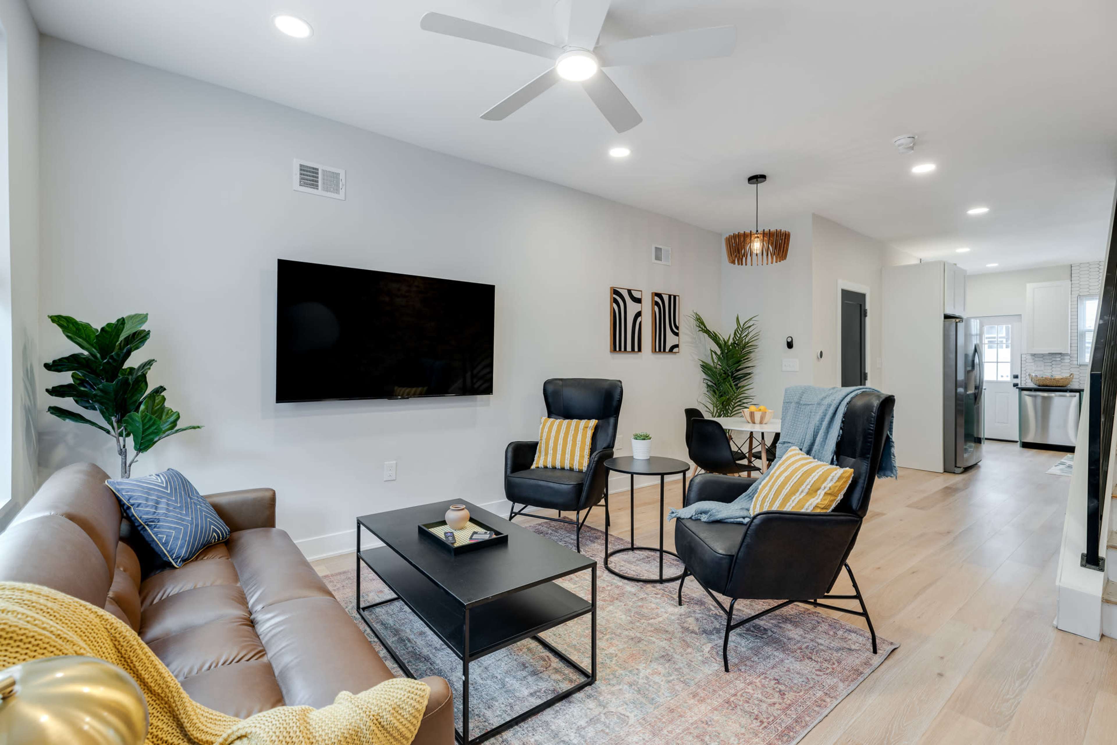 A modern living room features a brown leather sofa, two black chairs, a coffee table, and a mounted television, with decorative plants and artwork on the walls.