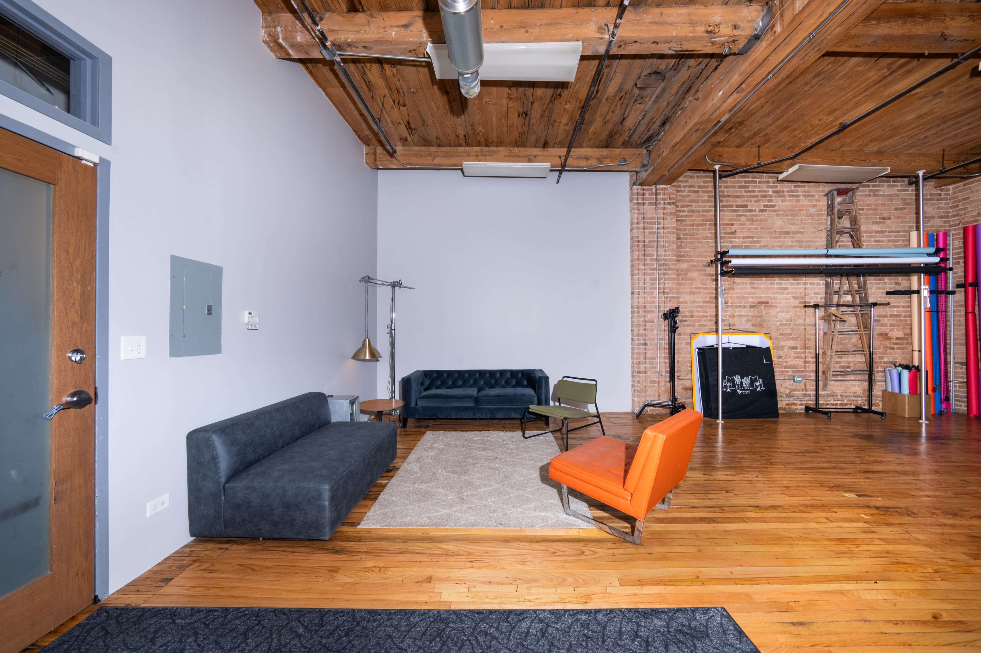 The image shows a spacious room with wooden floors, featuring a seating area with a gray sofa, an orange chair, and a large area rug against a backdrop of exposed brick walls and industrial elements.