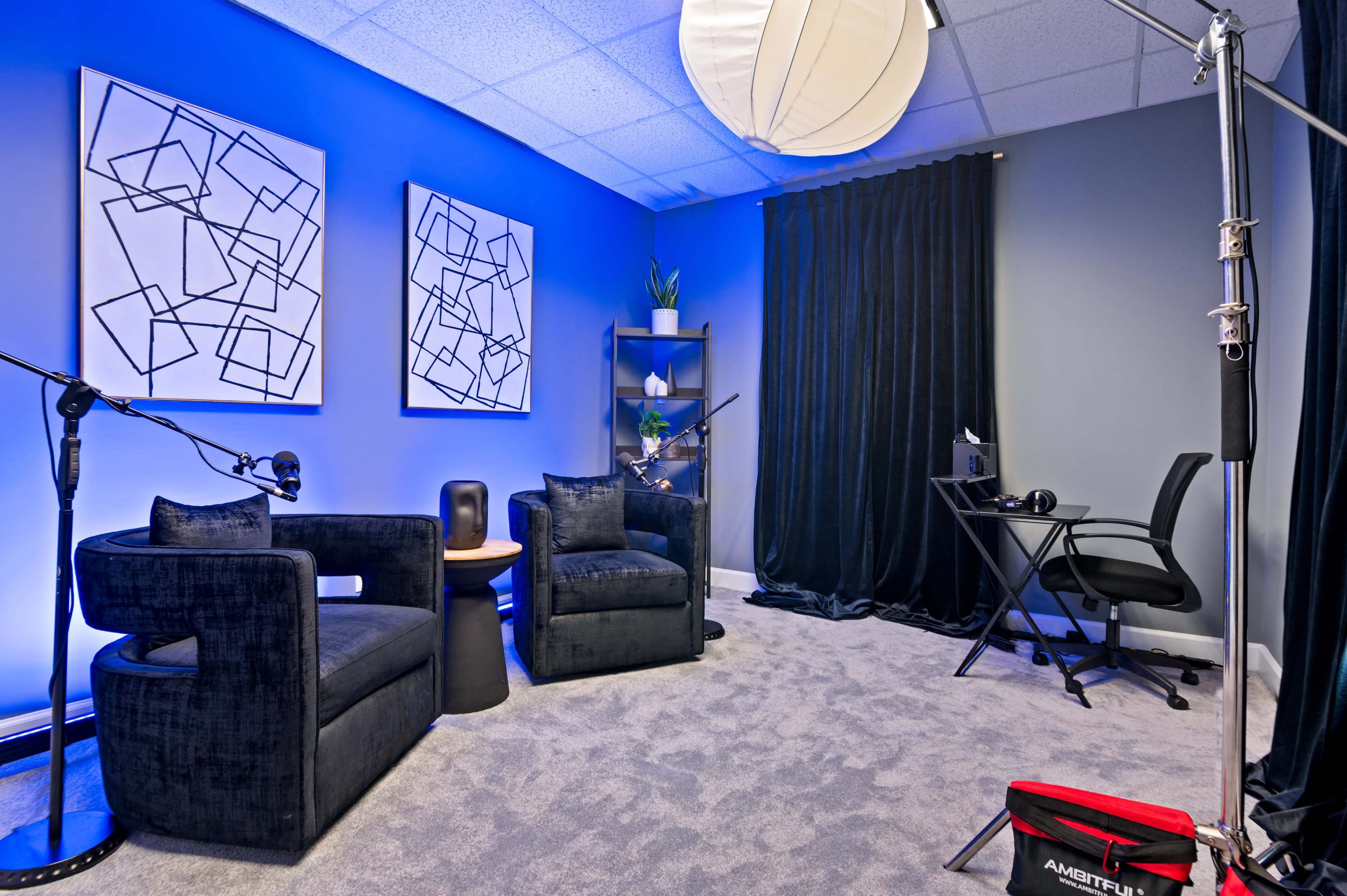 A small, modern room features two dark chairs, abstract artwork on the walls, a black desk with a computer setup, and a large softbox light.