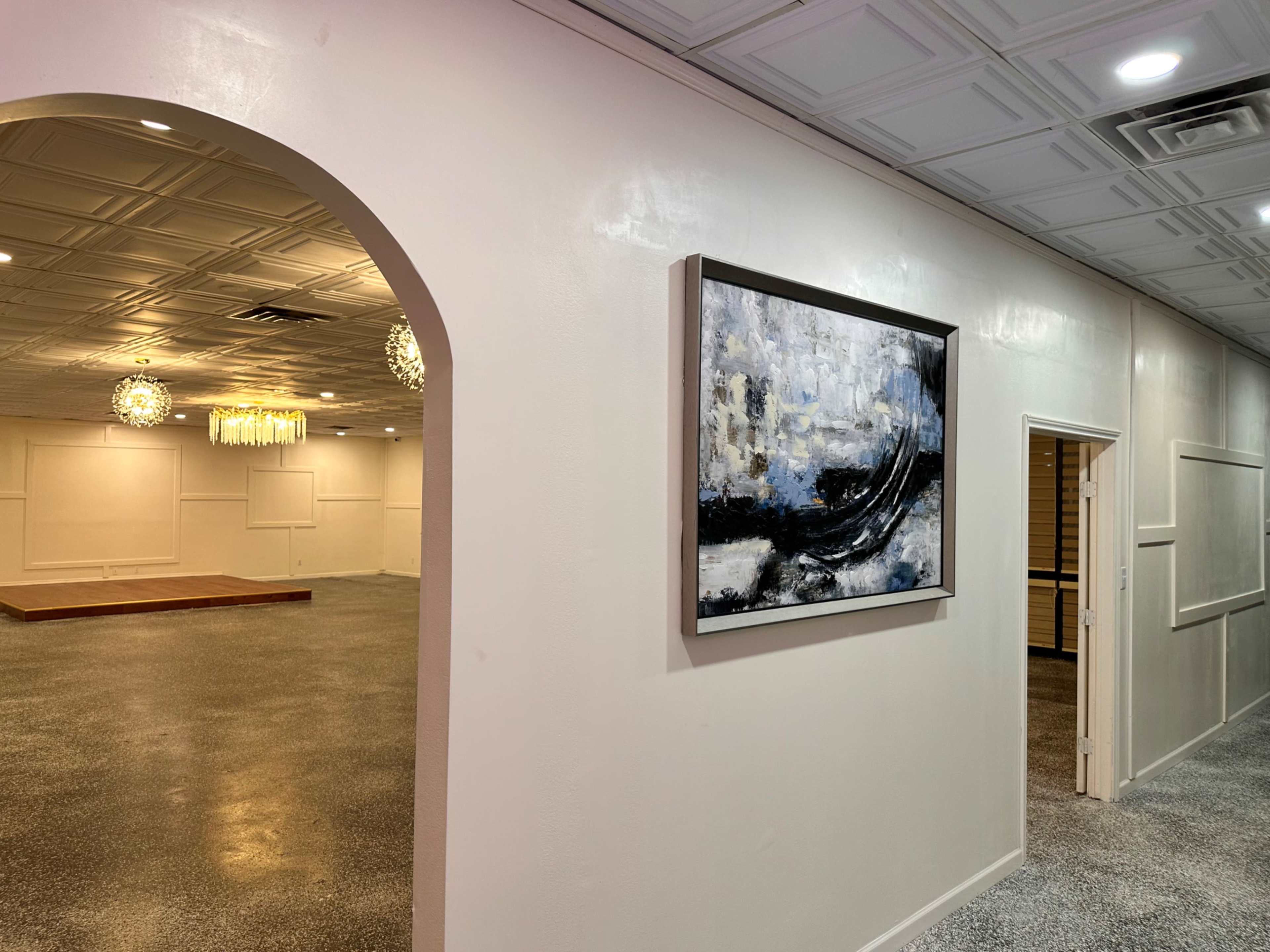 A corridor leading to a large, empty event space, with a painting hanging on the wall and decorative lighting in the distance.