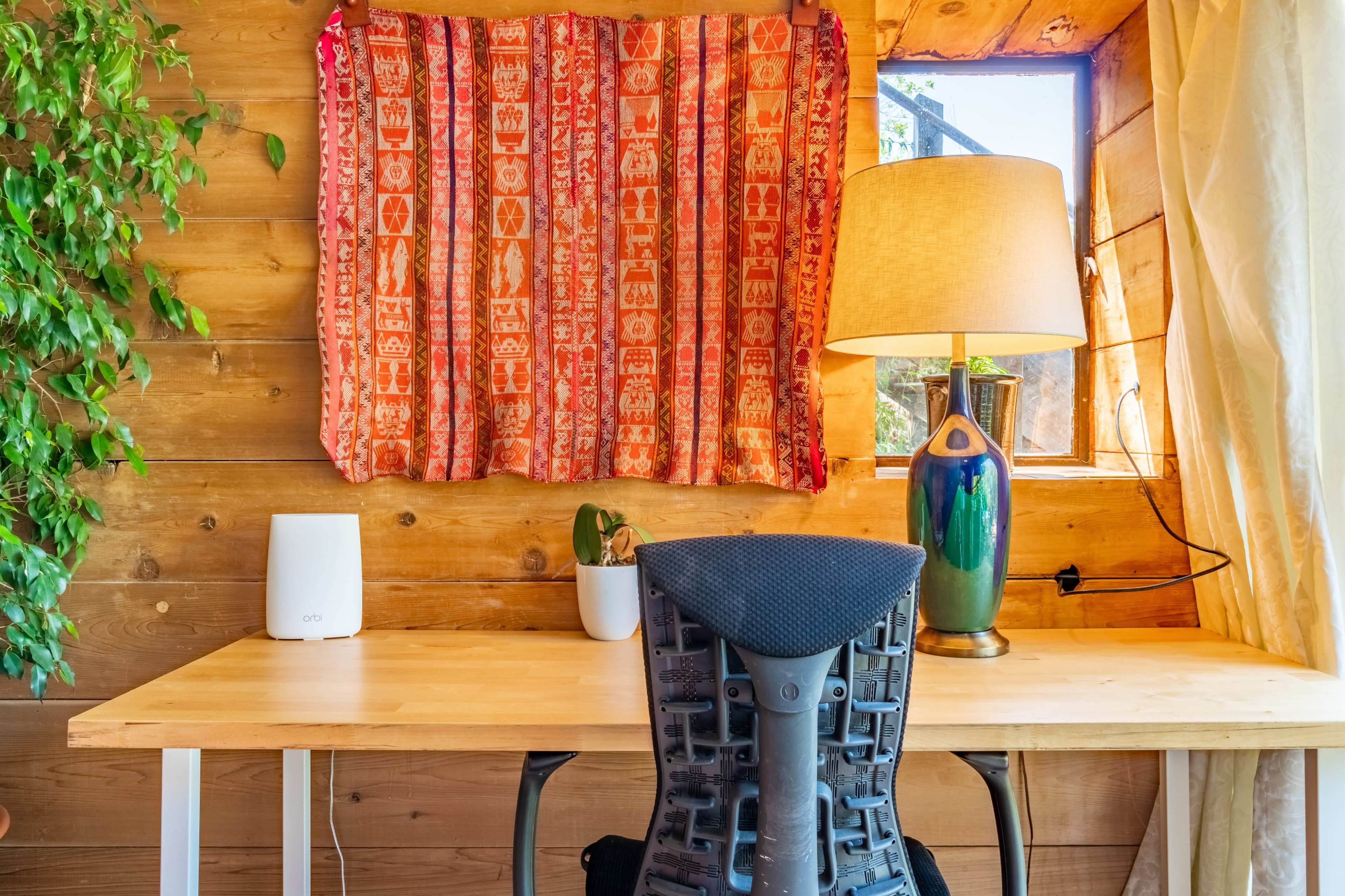 A wooden workspace featuring a desk with a chair, a decorative fabric hanging over a window, a lamp, and a small plant.