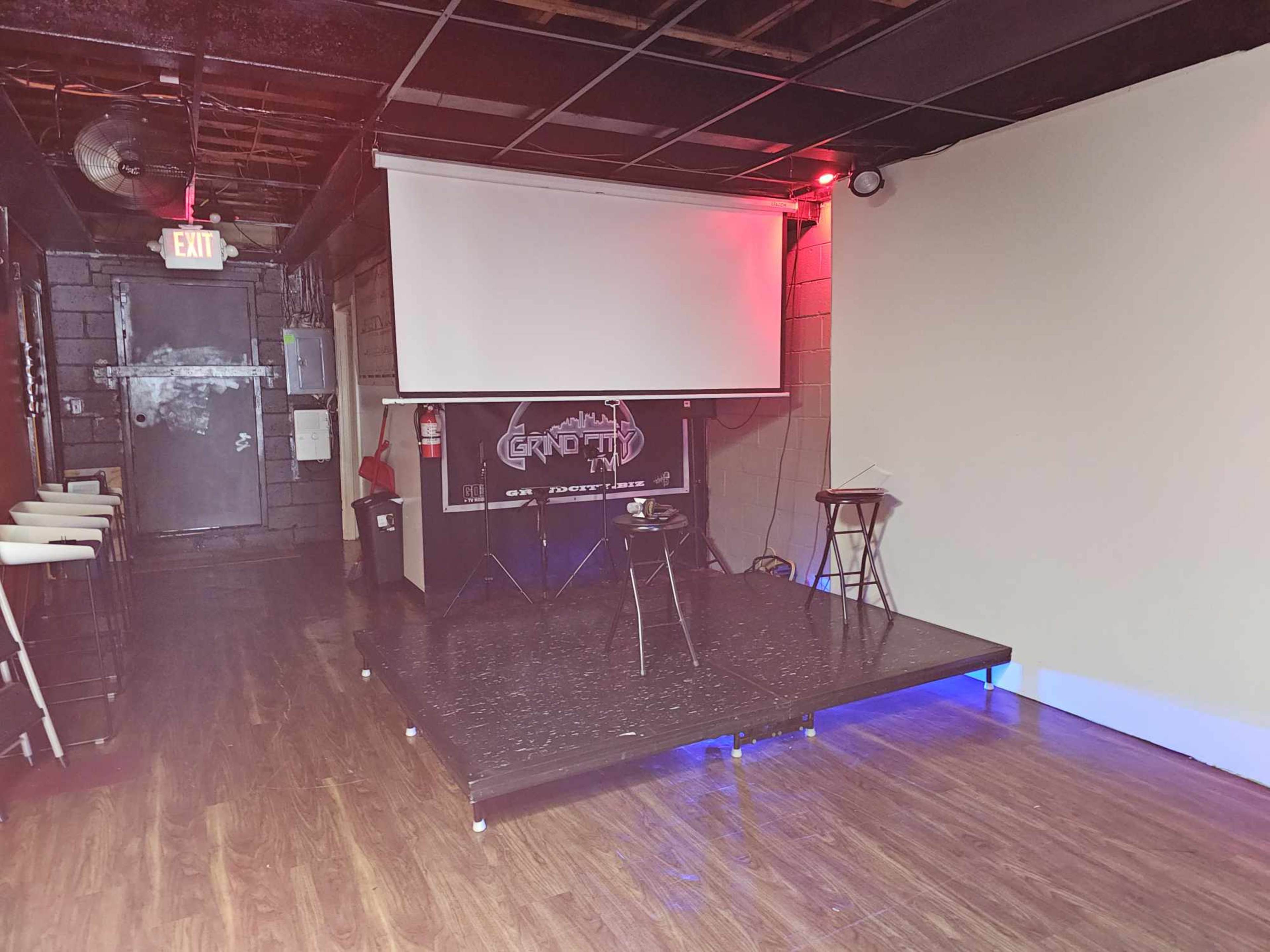 A dimly lit room with a stage featuring two stools, a projector screen, and a bar area in the background.