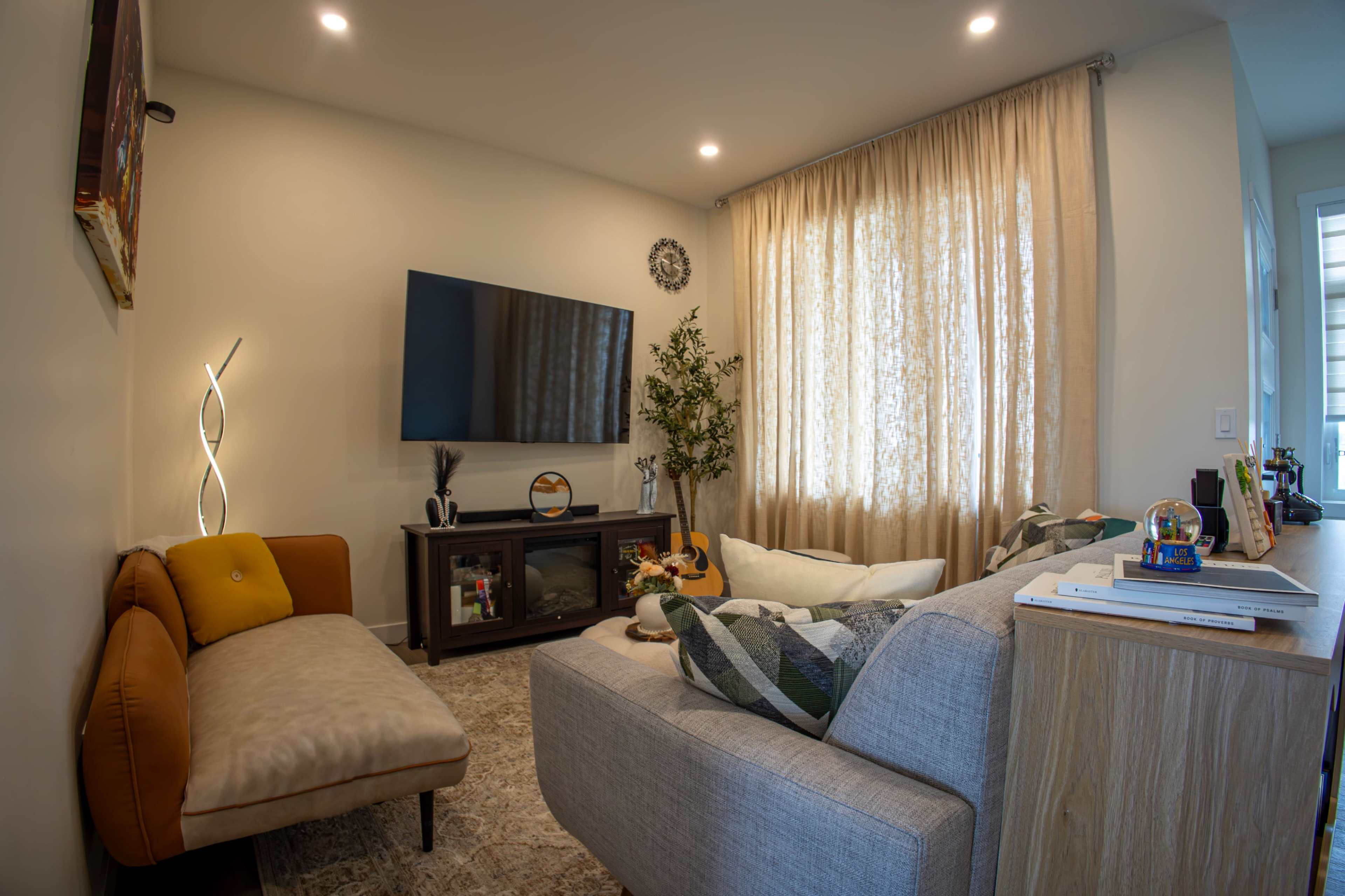 The image shows a cozy living room featuring a large television mounted on the wall, a neutral-toned sectional sofa, a chaise lounge, and a window with light curtains.