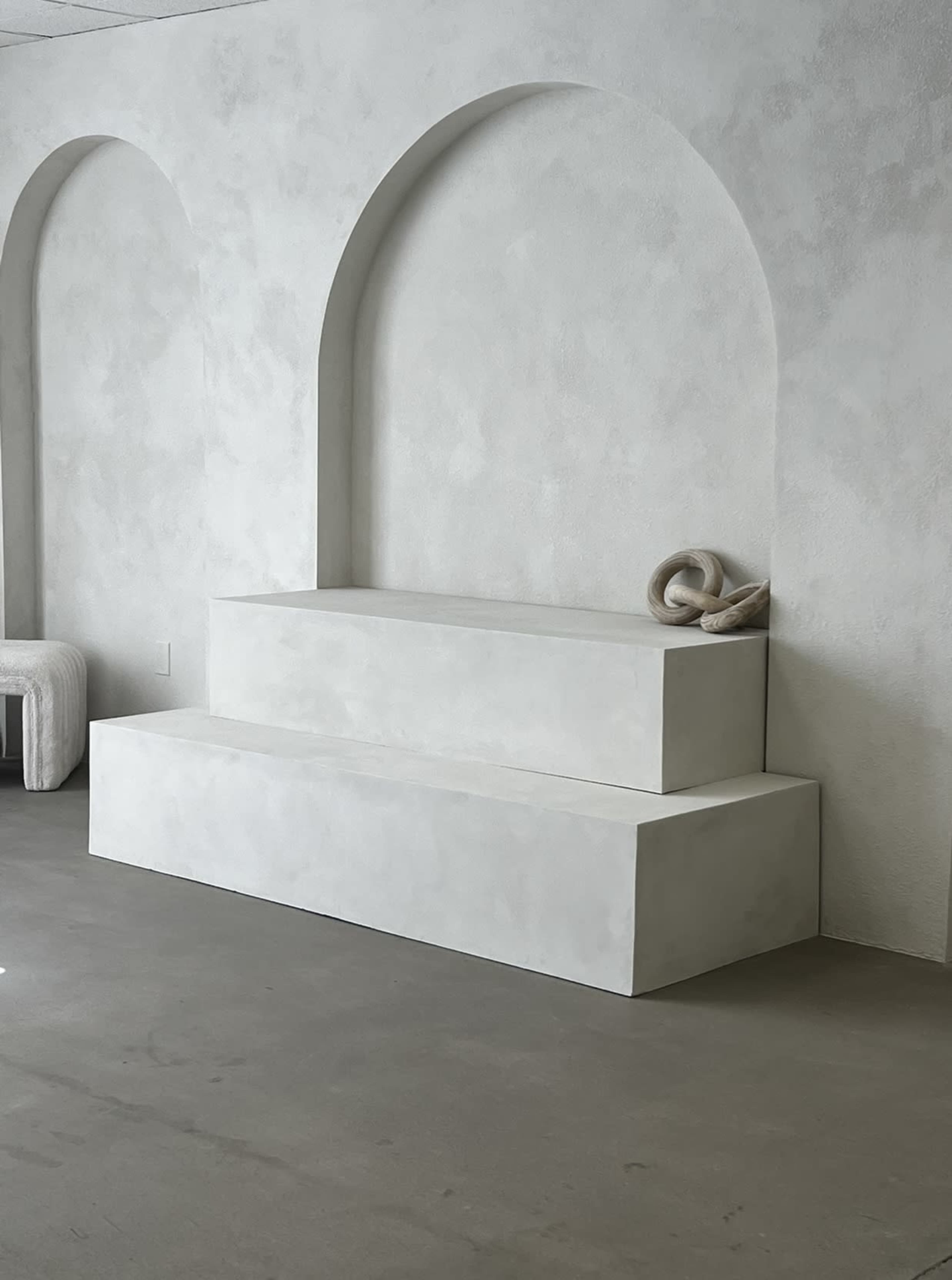 A minimalist interior features two stacked, rectangular concrete platforms beneath arched wall recesses.