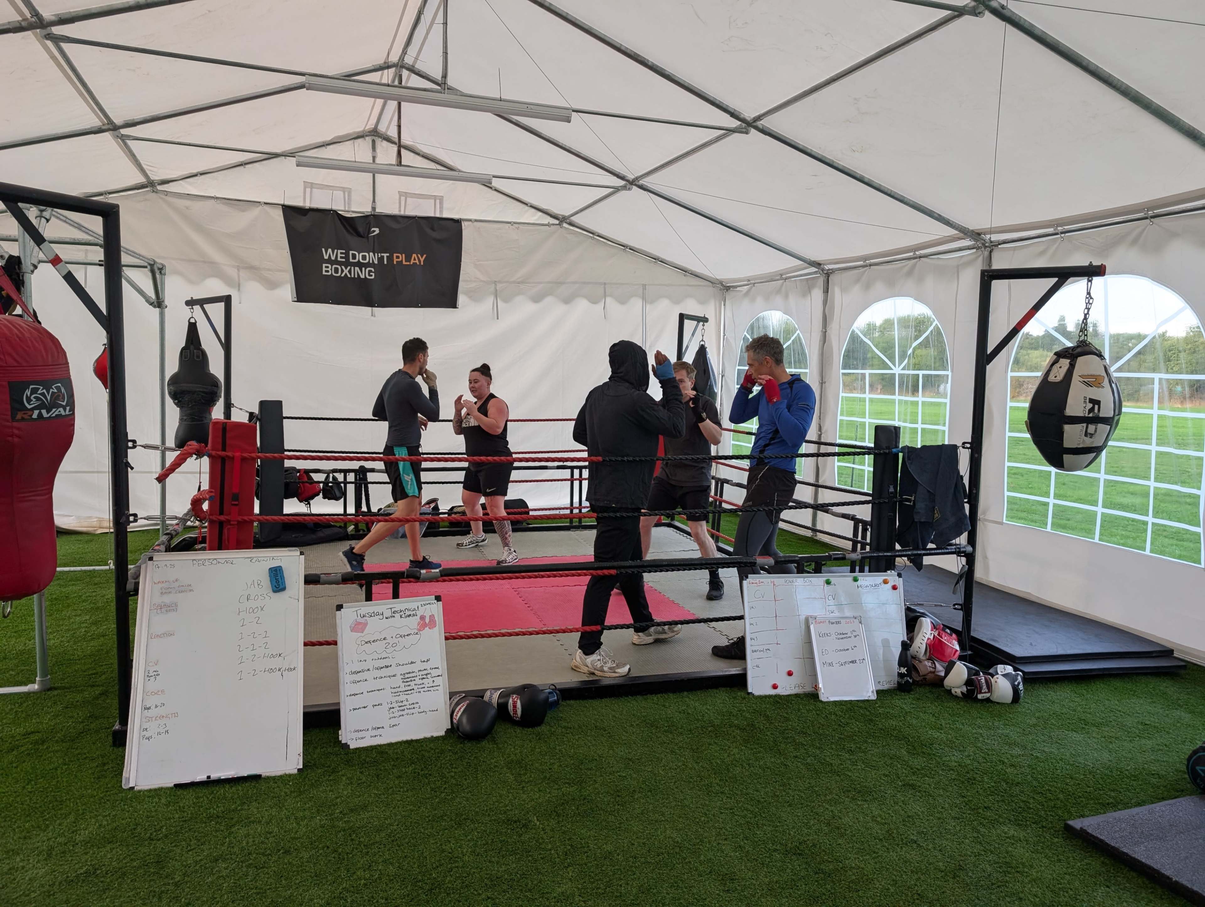 A group of individuals is training in a boxing ring inside a large tent with equipment and notes visible on the floor.