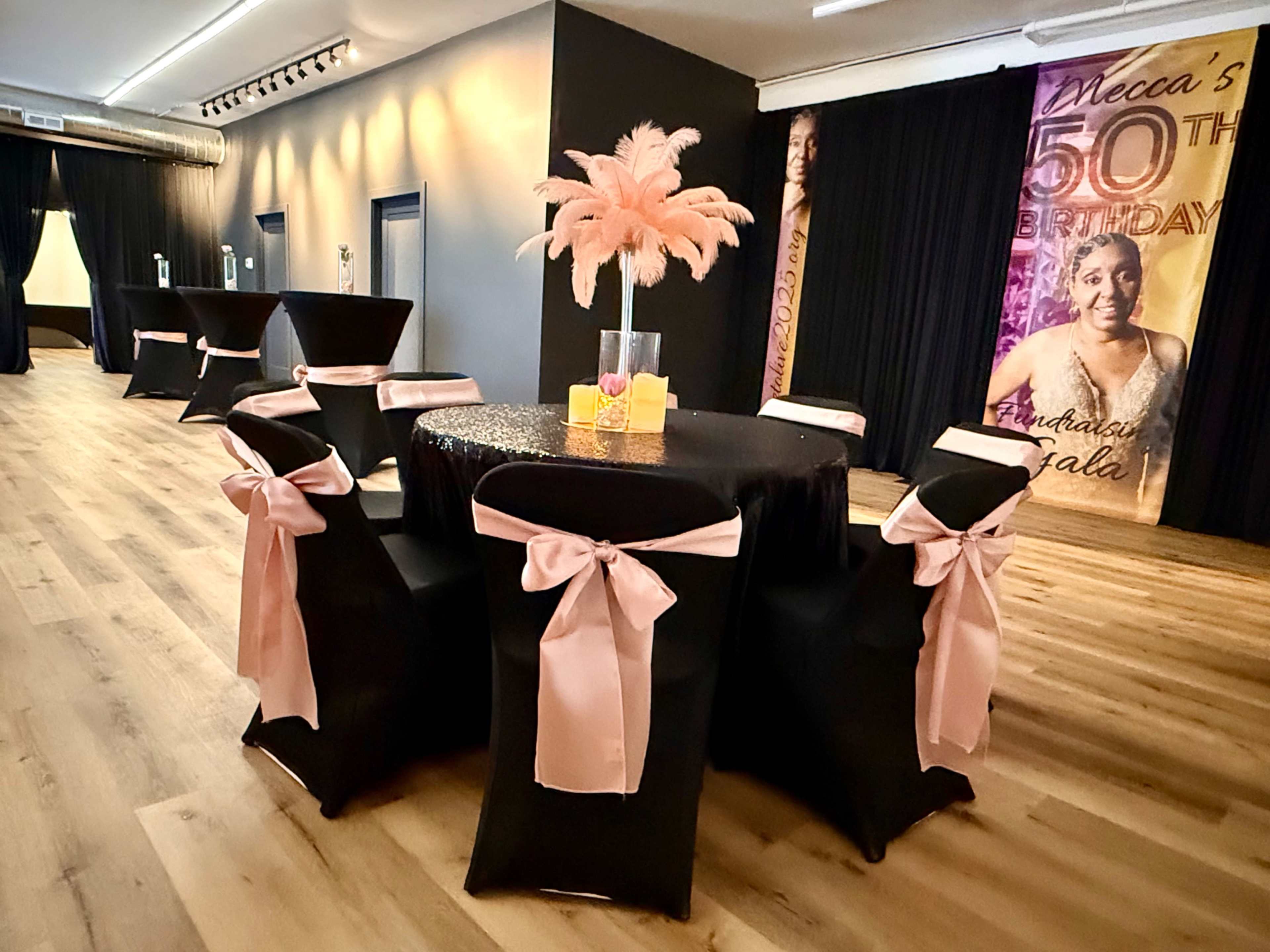 A beautifully decorated event space features a central table with a black tablecloth and pink accents, surrounded by chairs, with a backdrop celebrating a 50th birthday.