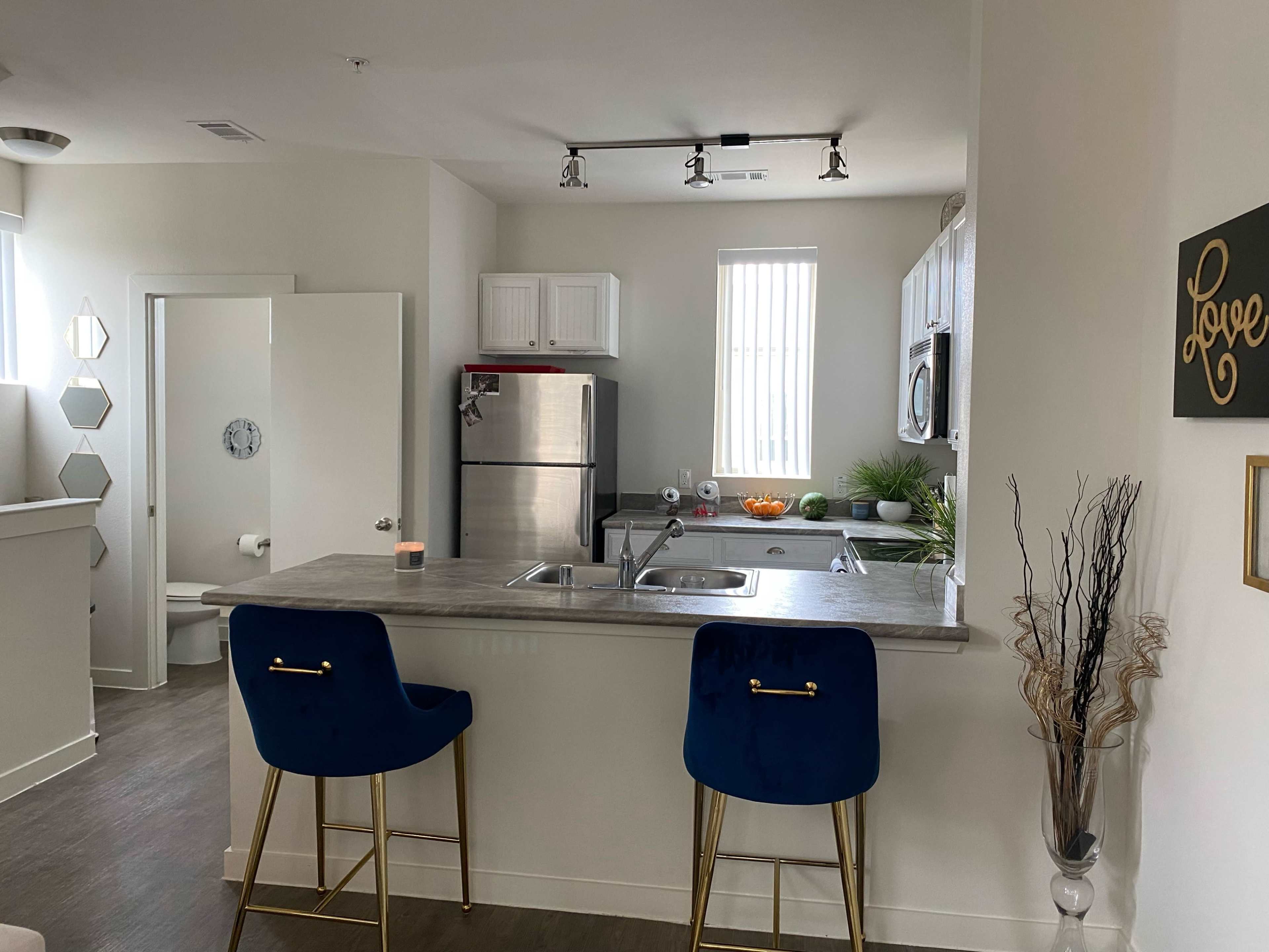 A modern kitchen with a small bar area featuring two blue stools, stainless steel appliances, and minimalist decor.