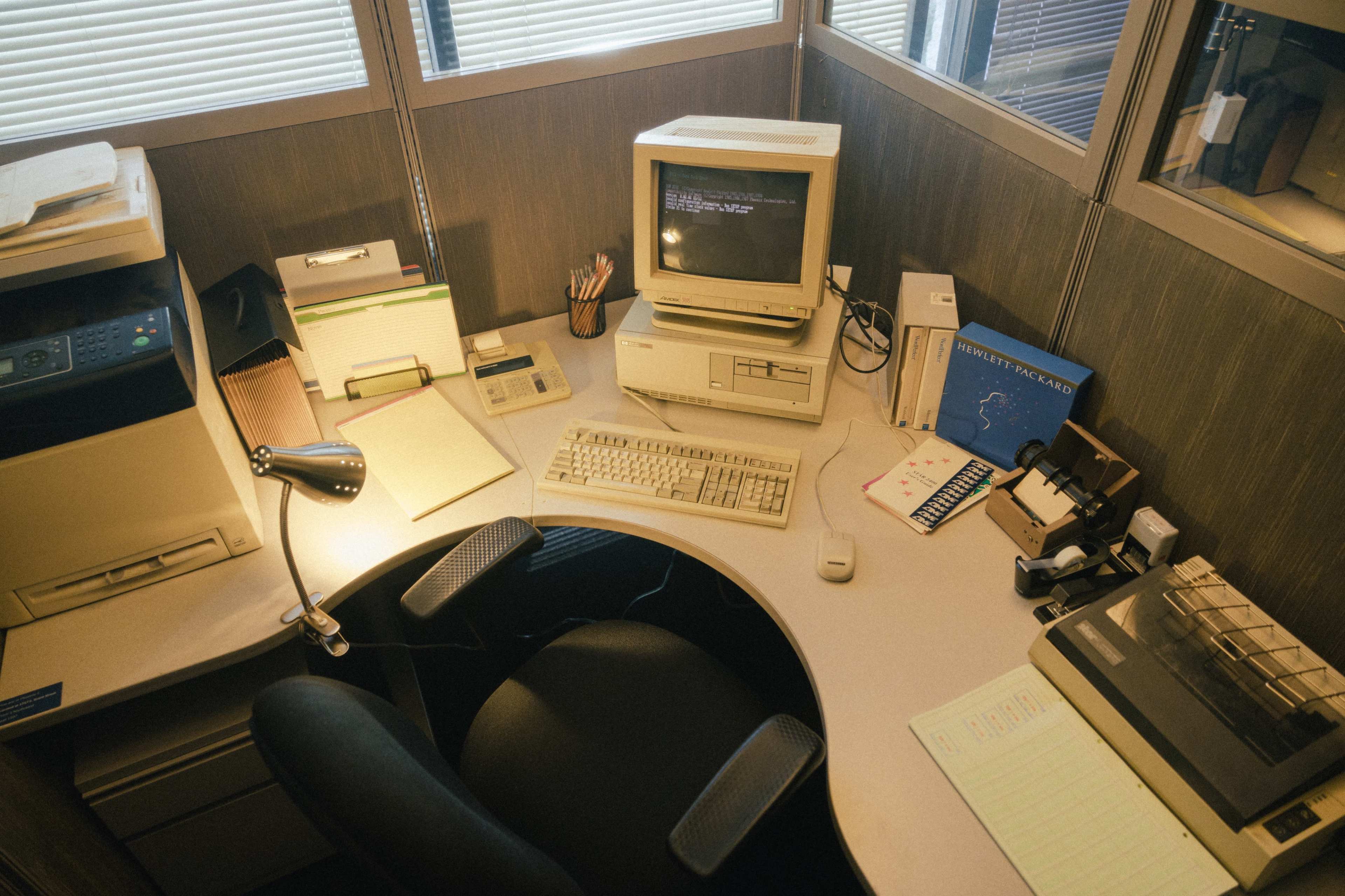 An overhead view of a cubicle office space featuring an old computer, printer, desk lamp, and various office supplies neatly arranged on a desk.