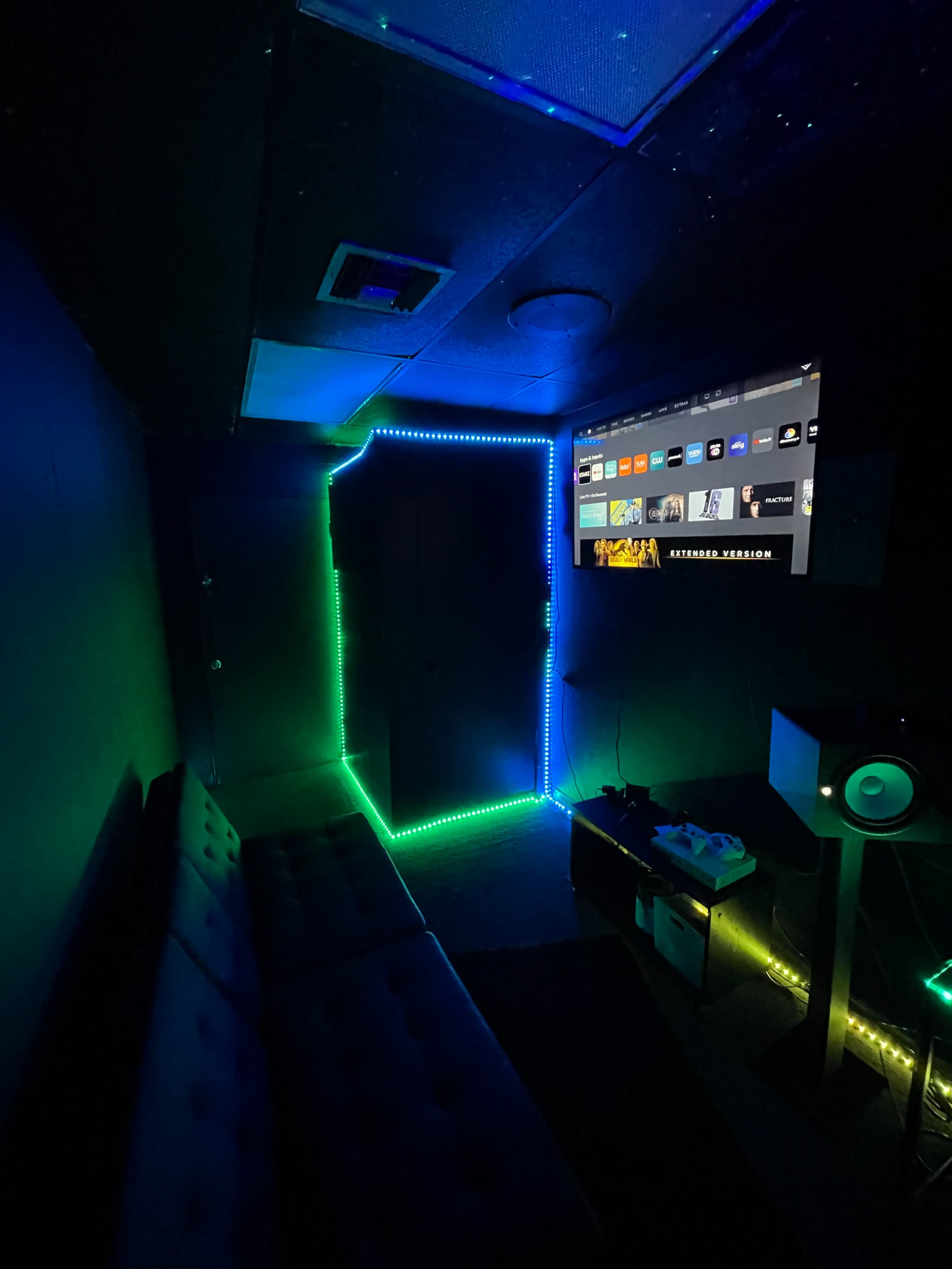 A dimly lit room features a couch, an LED-lit doorway, and a large wall-mounted screen displaying various media options.