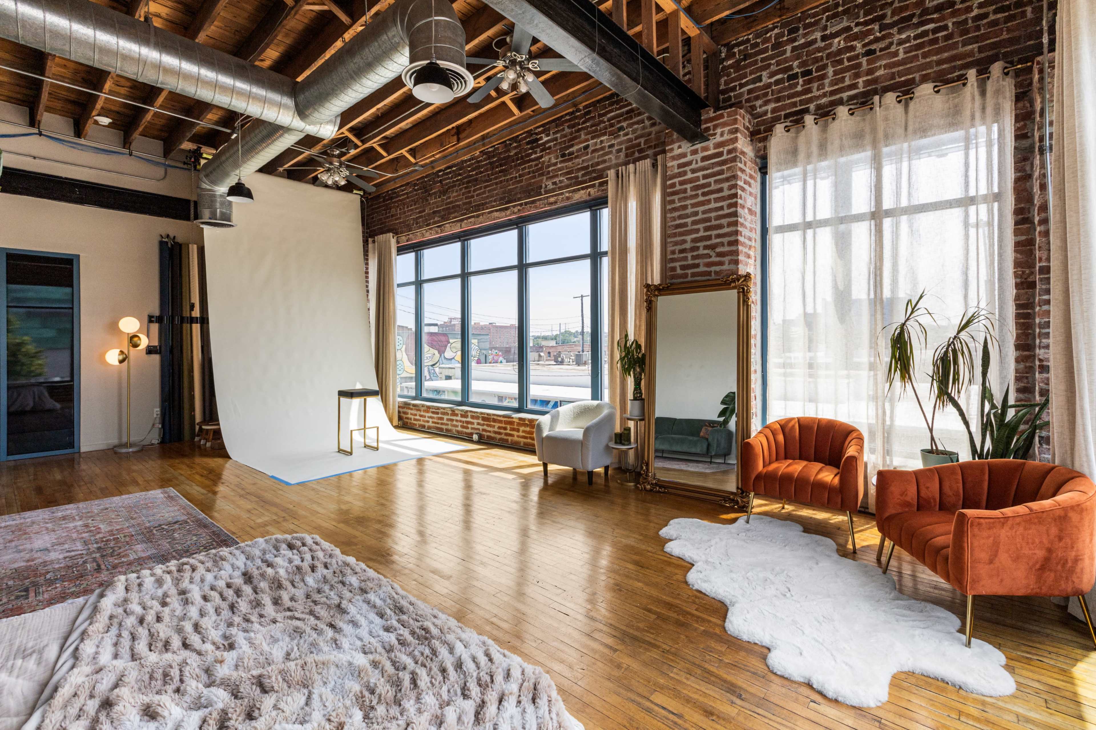 The image shows a spacious loft with exposed brick walls, hardwood floors, large windows, and a cozy seating area featuring two orange chairs and a decorative mirror.