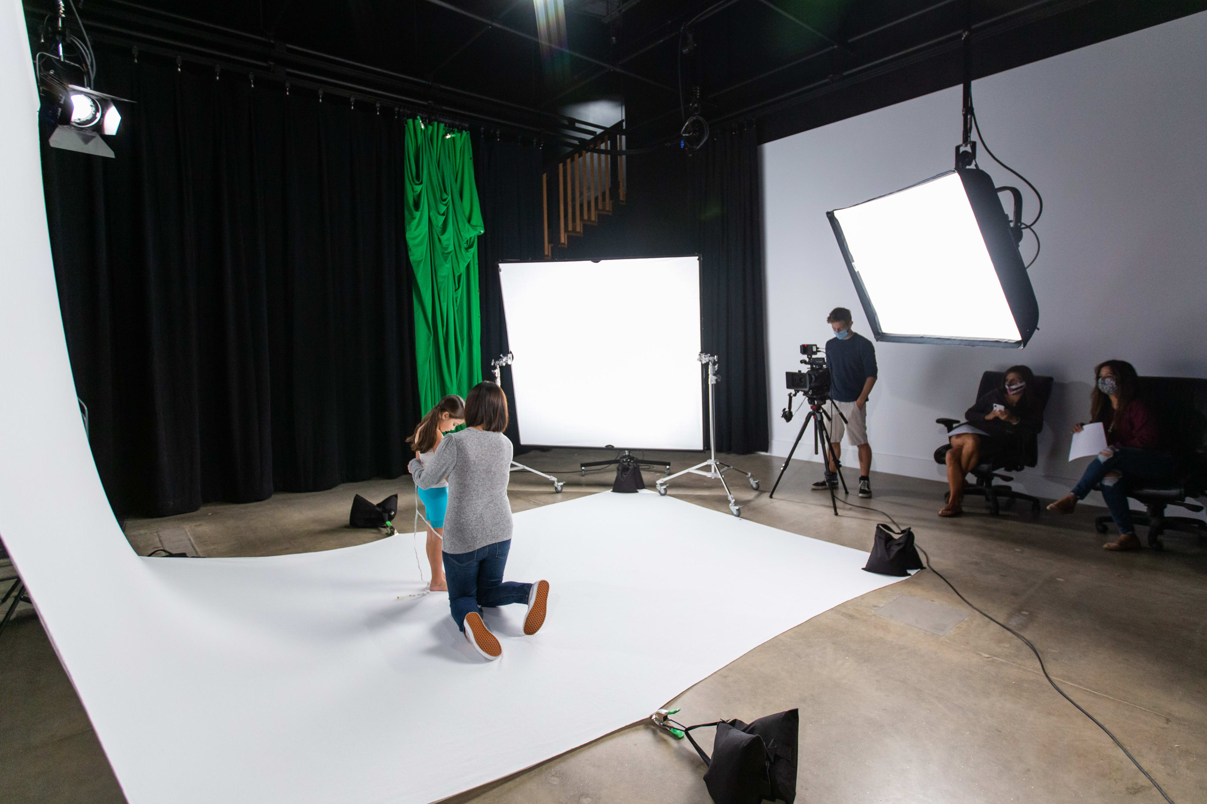 A child poses for a photo in a studio while a photographer adjusts the scene and crew members observe from a distance.