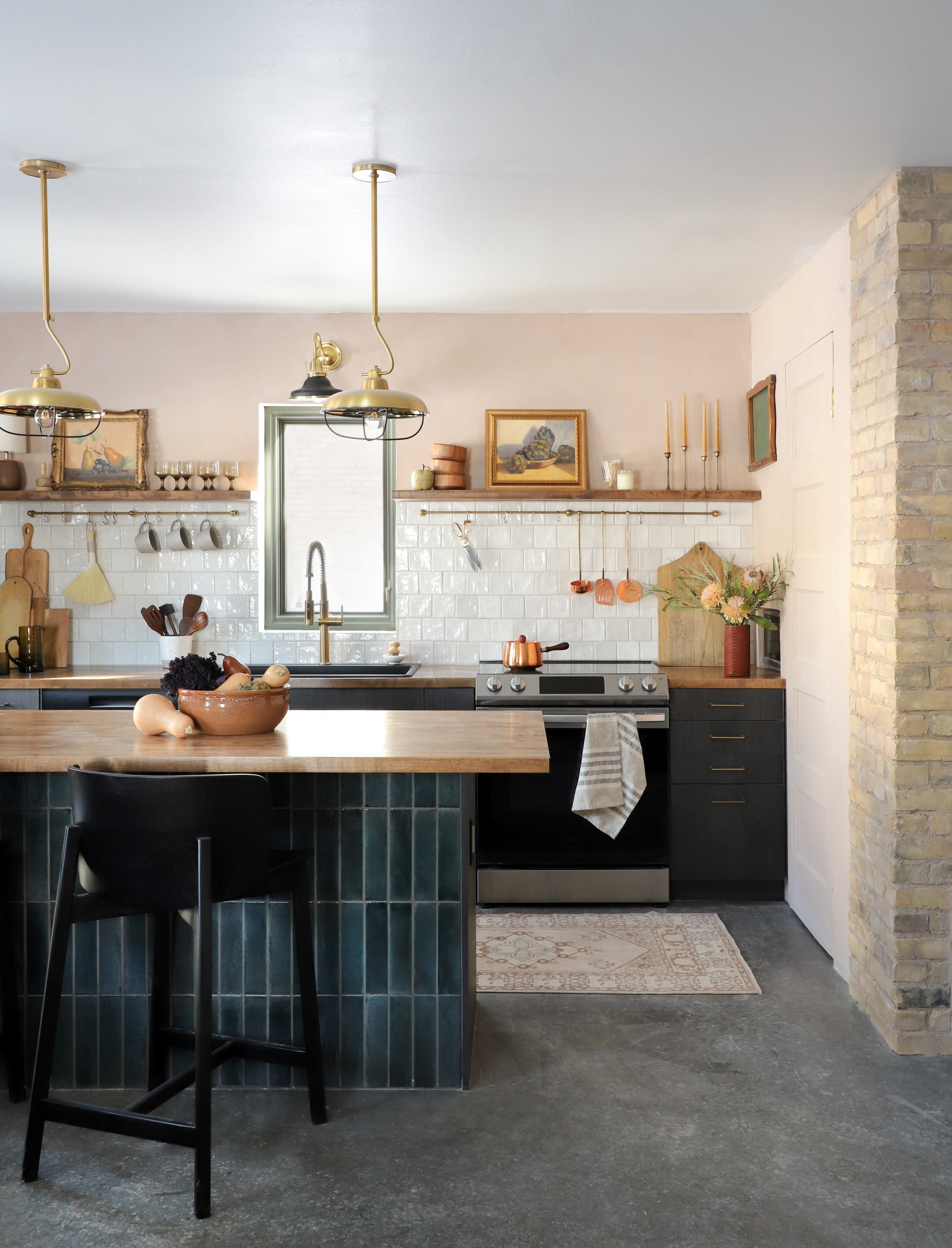 A modern kitchen features a central wooden island with bar stools, a stove against a tiled backsplash, and shelves displaying kitchenware and decor.