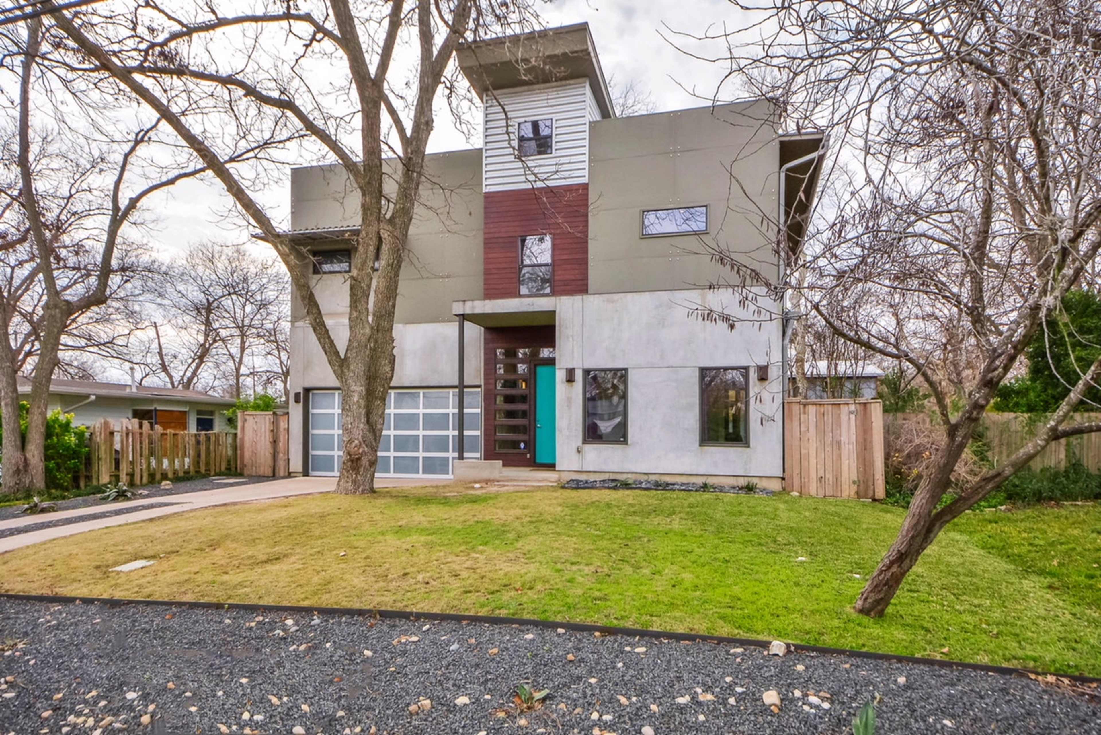 A modern three-story house with a mixture of concrete and wood paneling, surrounded by bare trees and a manicured lawn.