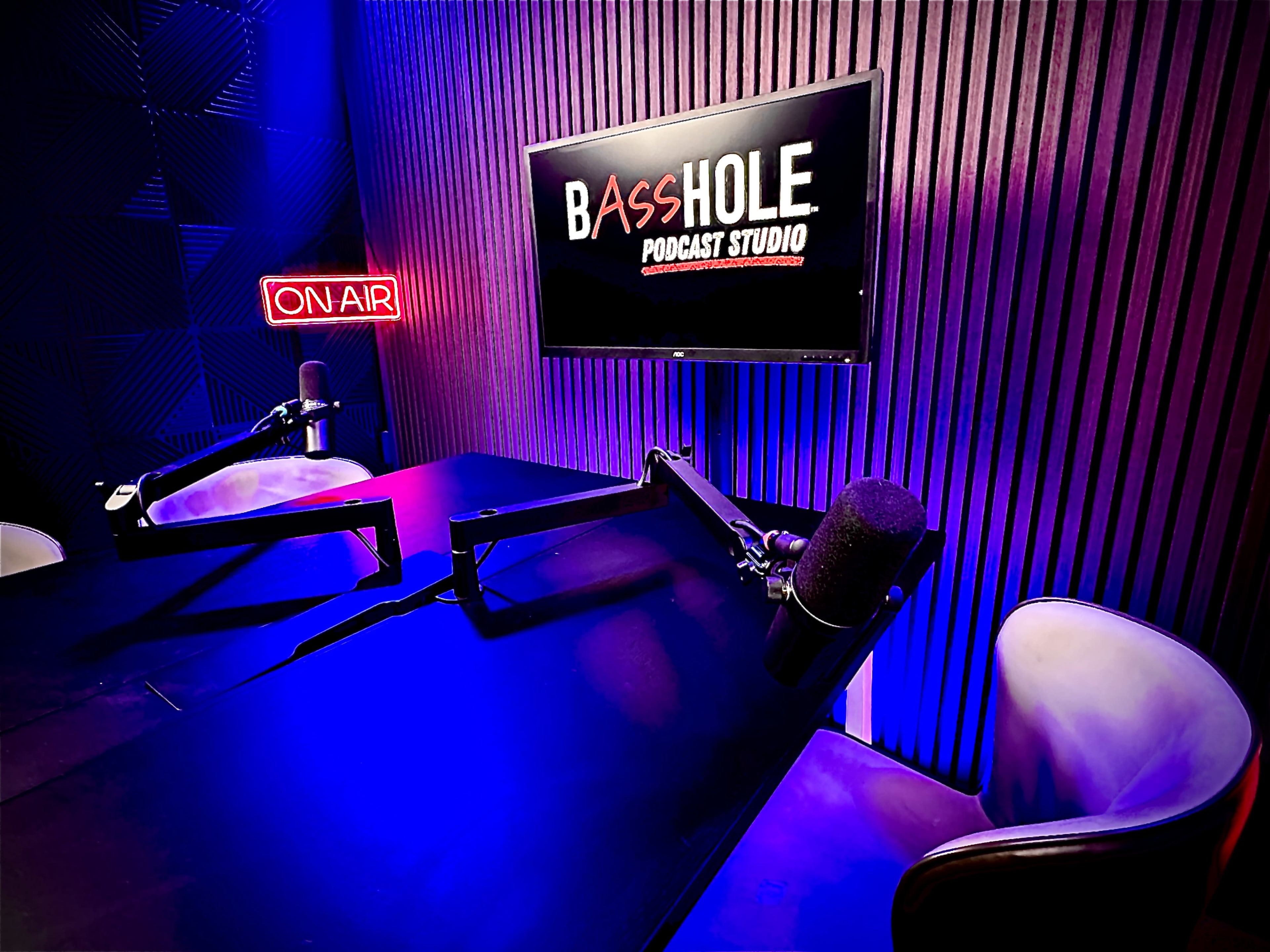 The image shows a podcast studio featuring a dark table, two microphones, and a monitor displaying the words "BASSHOLE PODCAST STUDIO," alongside a glowing "ON AIR" sign.