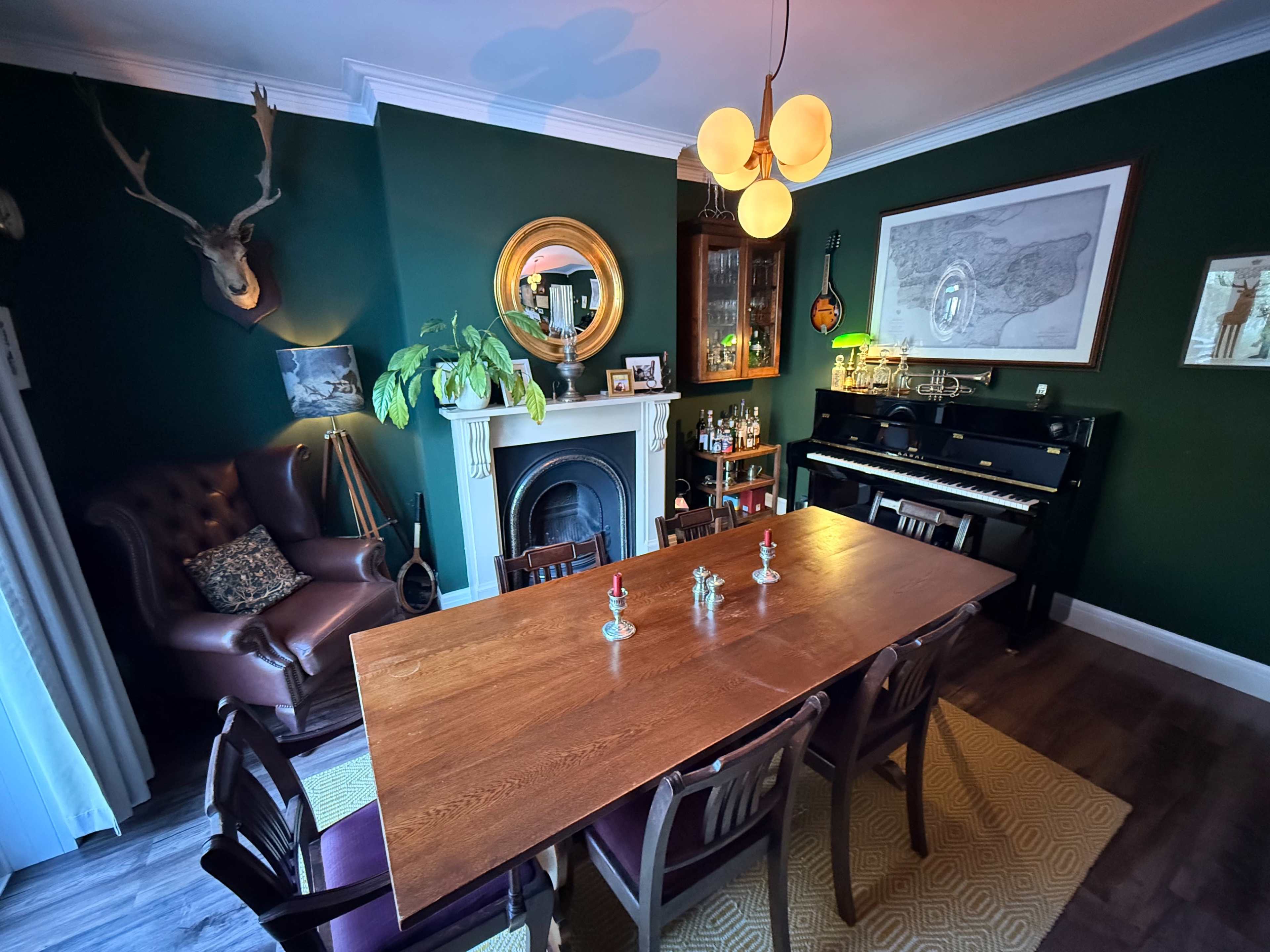 The image shows a dining room with a large wooden table, surrounded by chairs, a green wall adorned with a mirror and various decorations, and a piano in the corner.