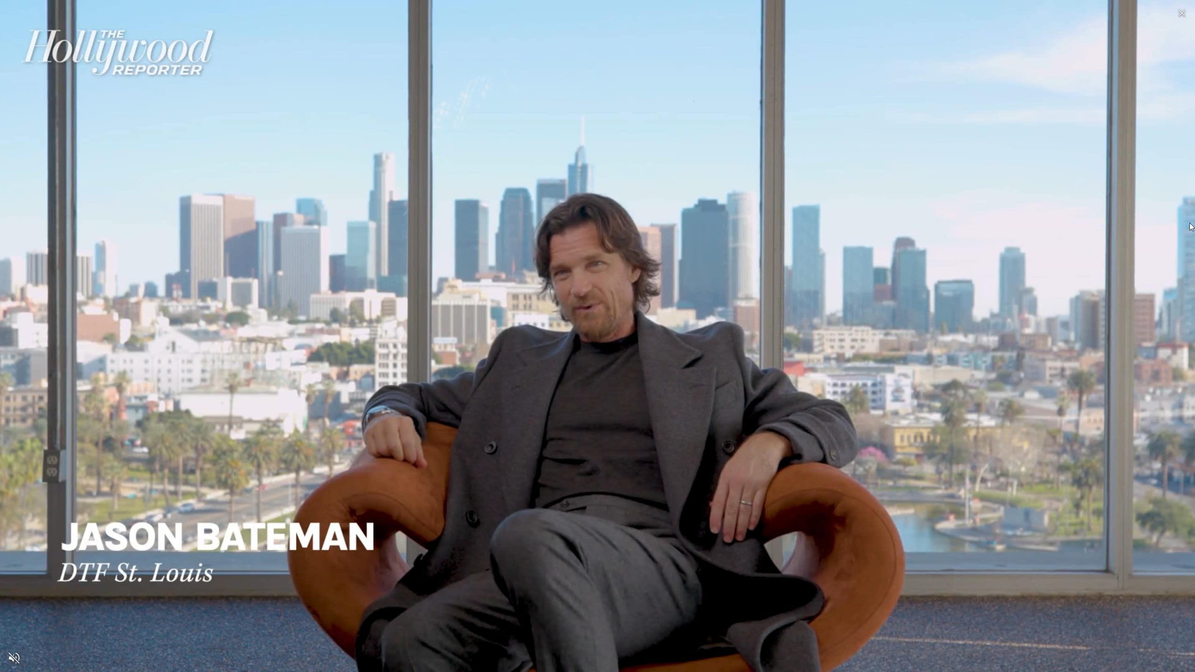 A man sits in a brown chair with a city skyline visible through large windows behind him.