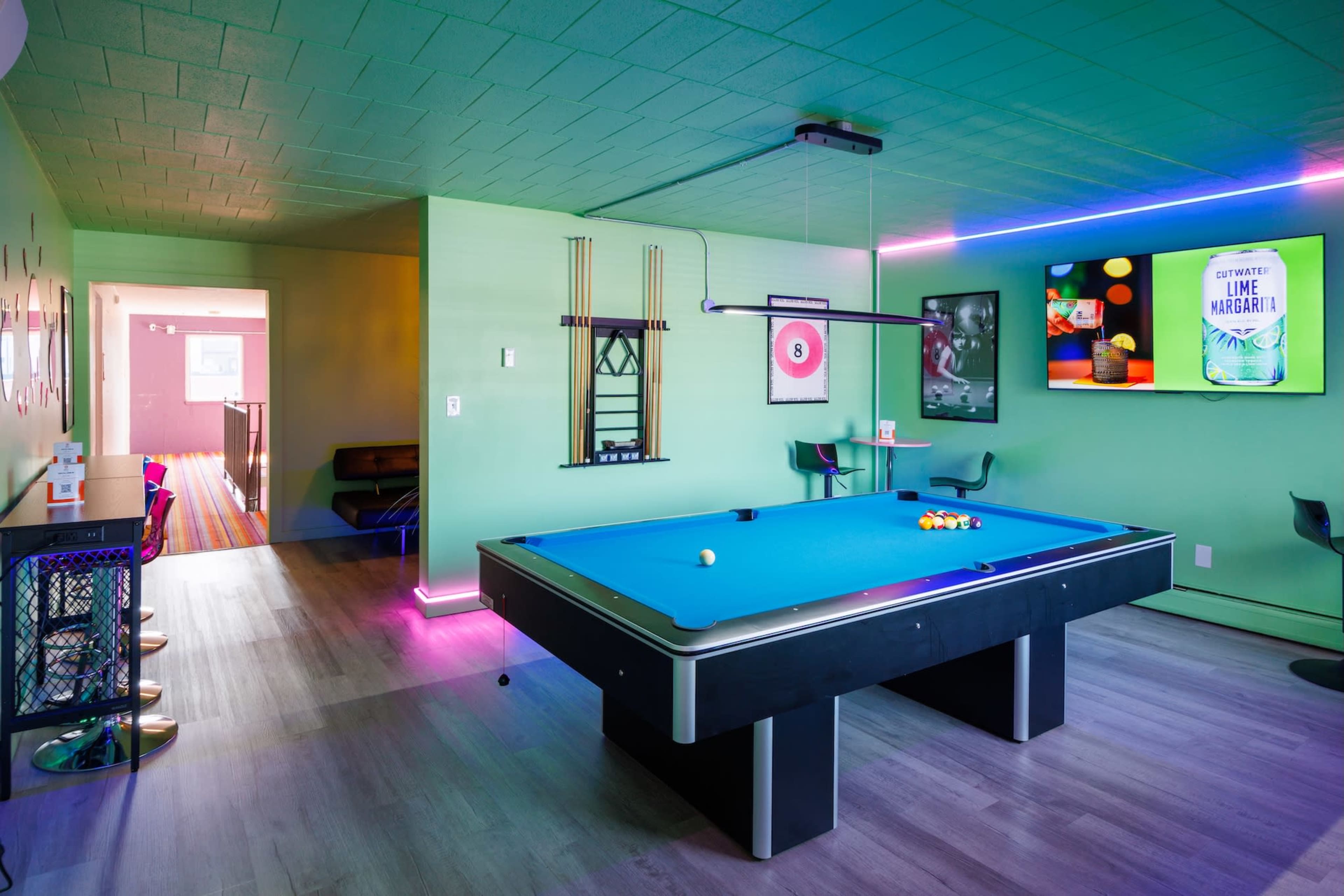The image shows a brightly colored game room featuring a pool table, modern bar stools, and neon lighting.