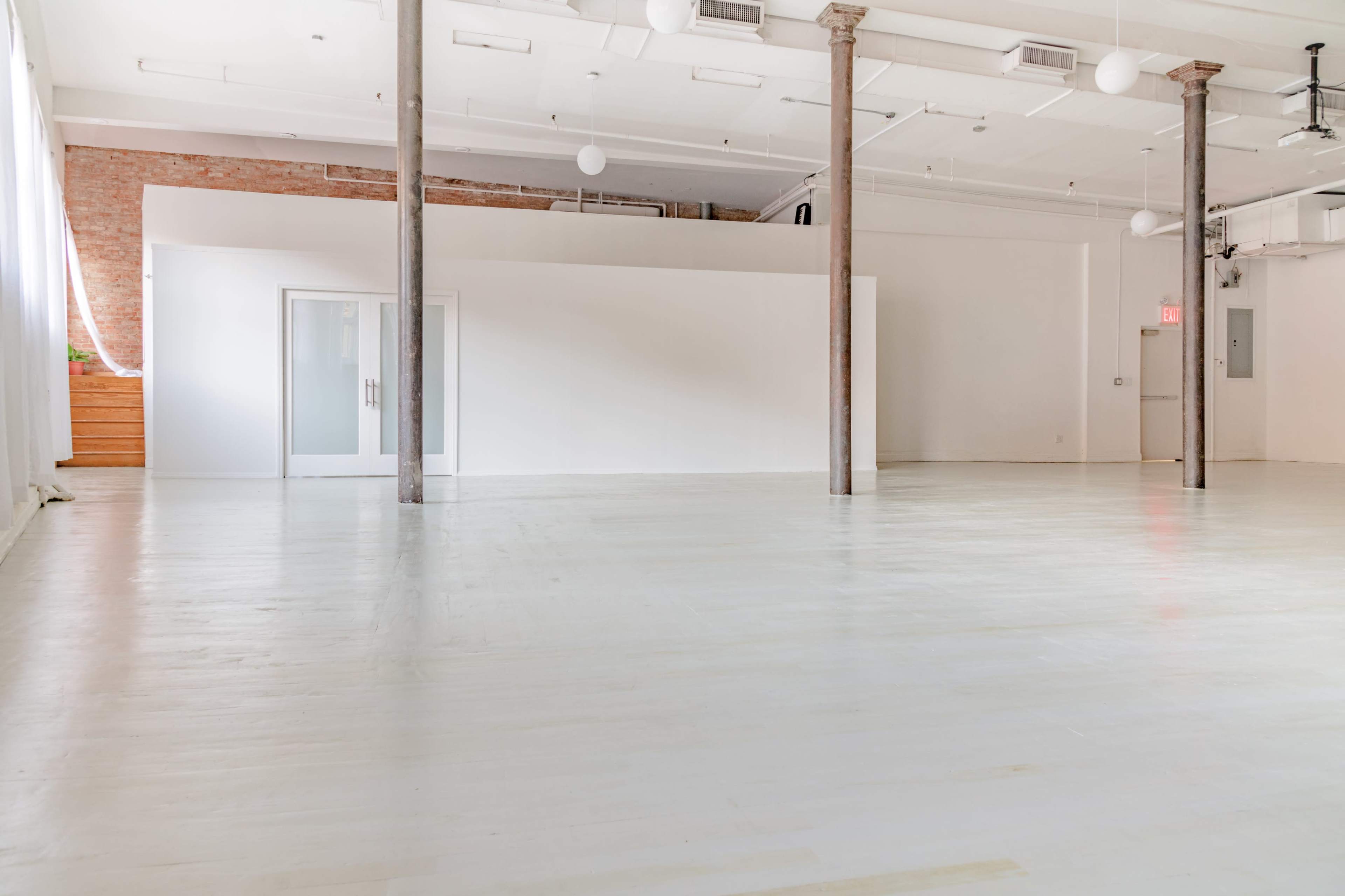 A spacious, empty room features light-colored wooden floors, white walls, and exposed columns, with large windows allowing natural light.