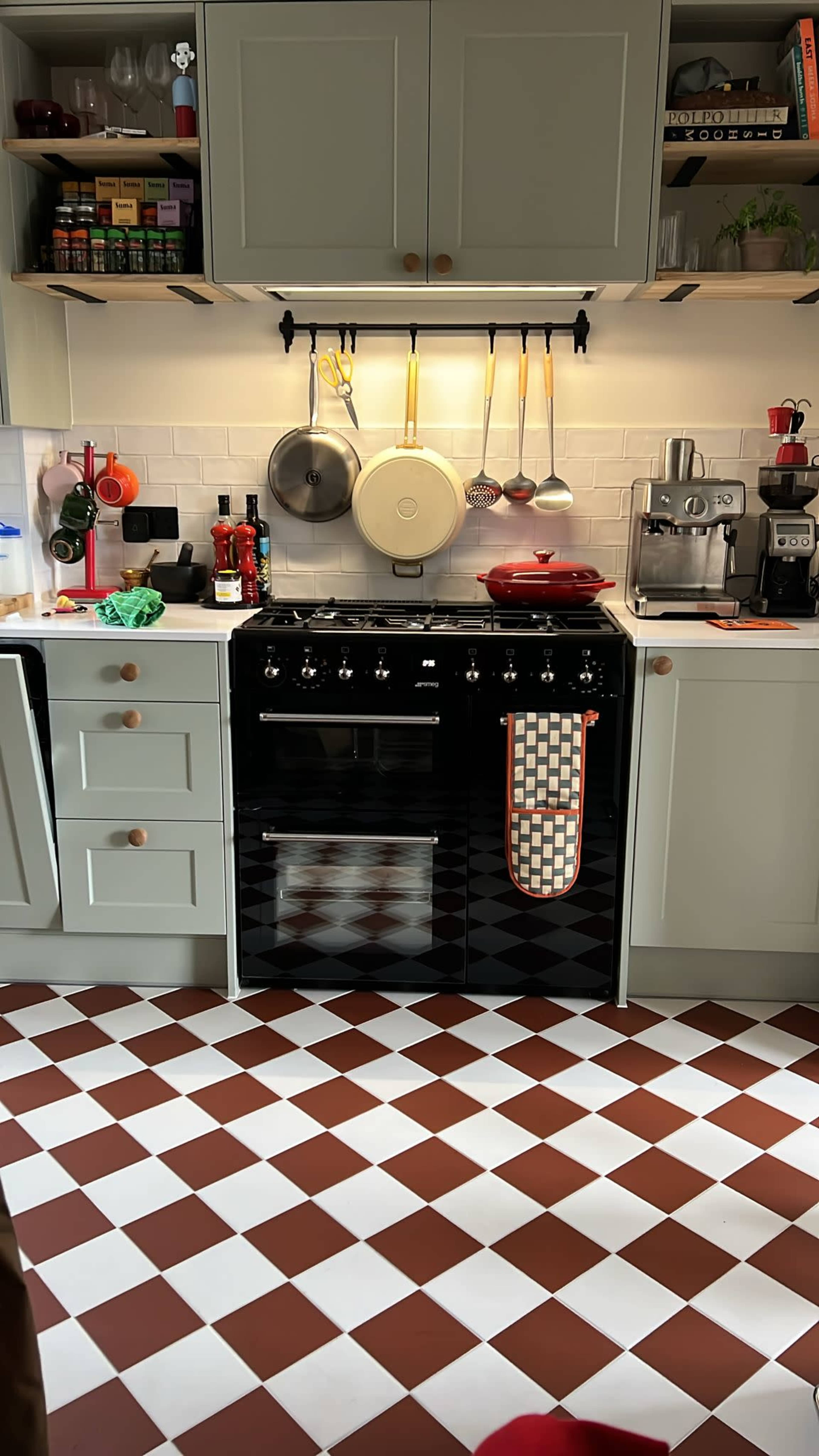The image shows a modern kitchen with a checkerboard floor, a black stove, stainless steel appliances, and neatly organized cookware hanging above the counter.