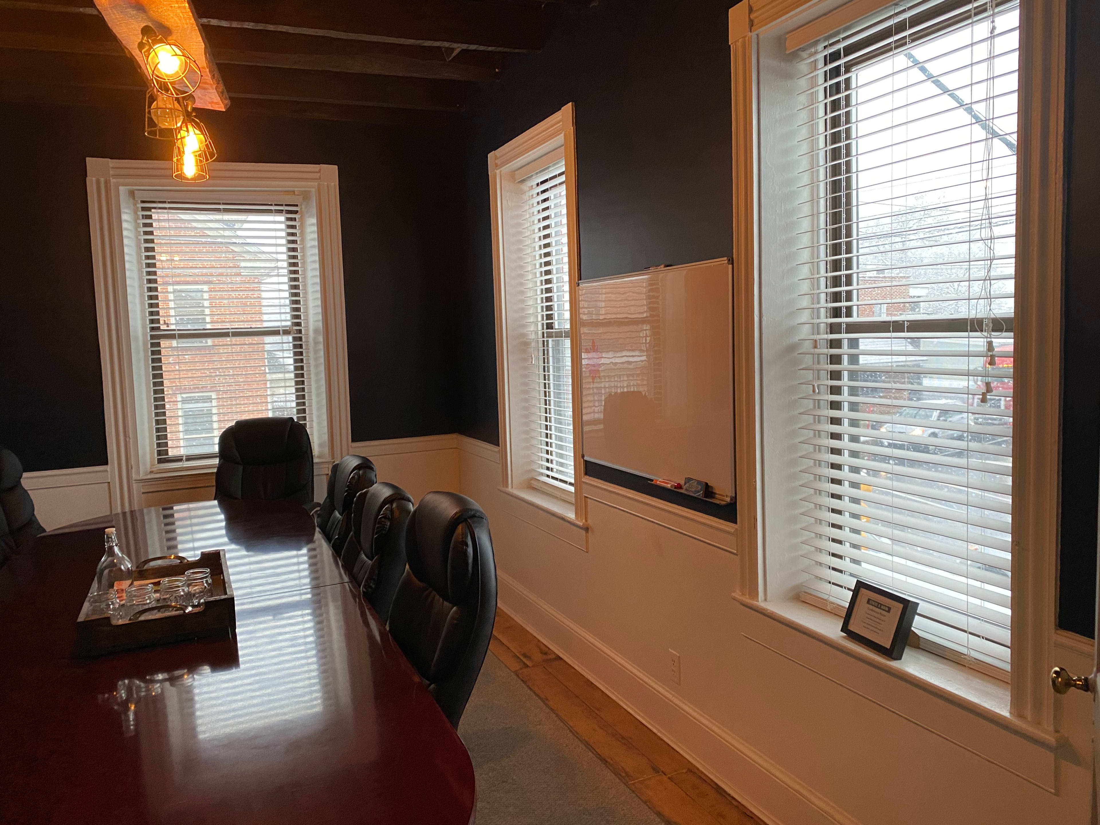 The image shows a conference room with a long wooden table, black leather chairs, and two large windows with blinds, featuring a whiteboard mounted on a wall.
