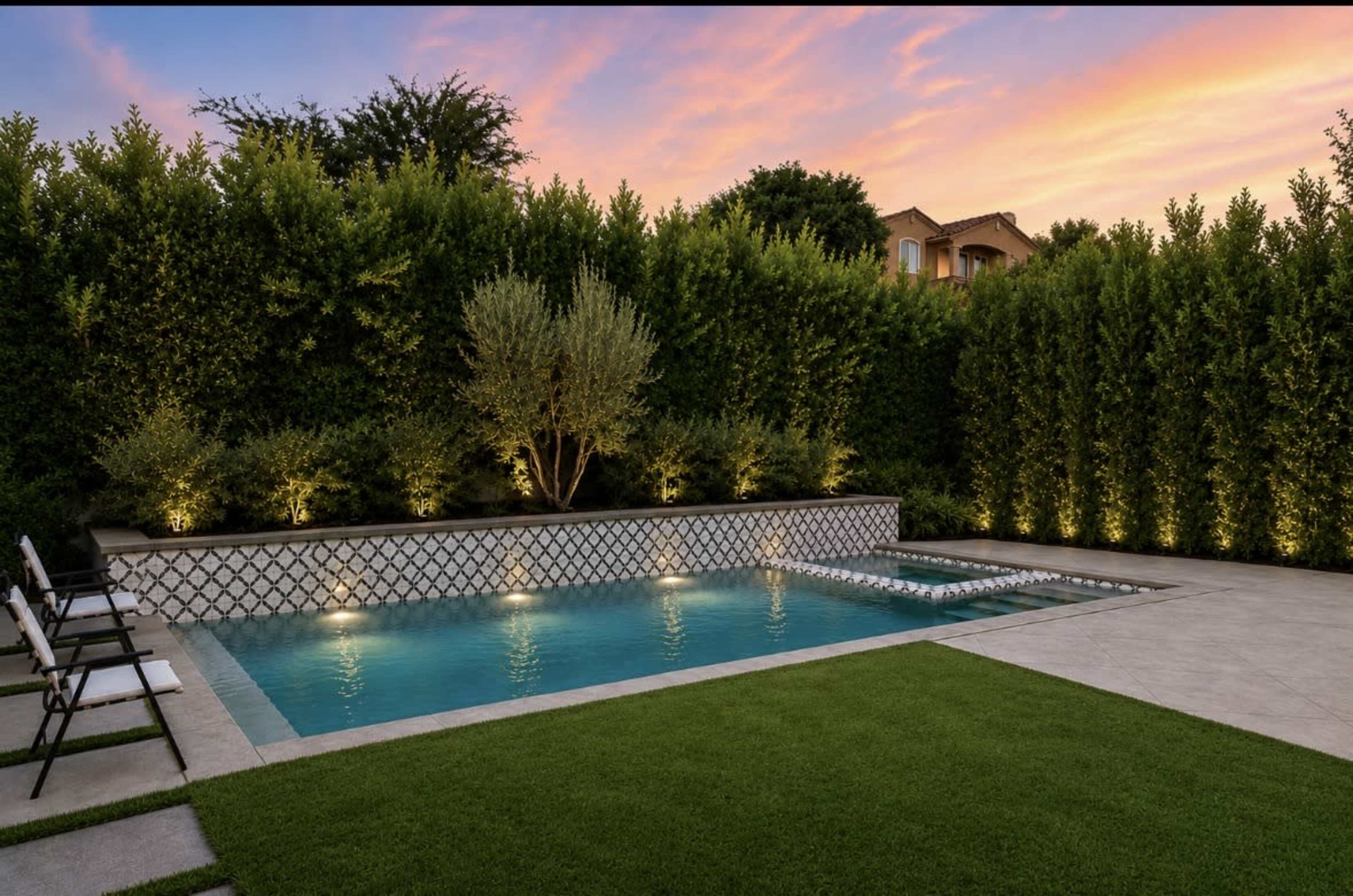 The image shows a modern swimming pool surrounded by lush greenery and a concrete deck, with lounge chairs positioned nearby and the sky illuminated by a sunset.