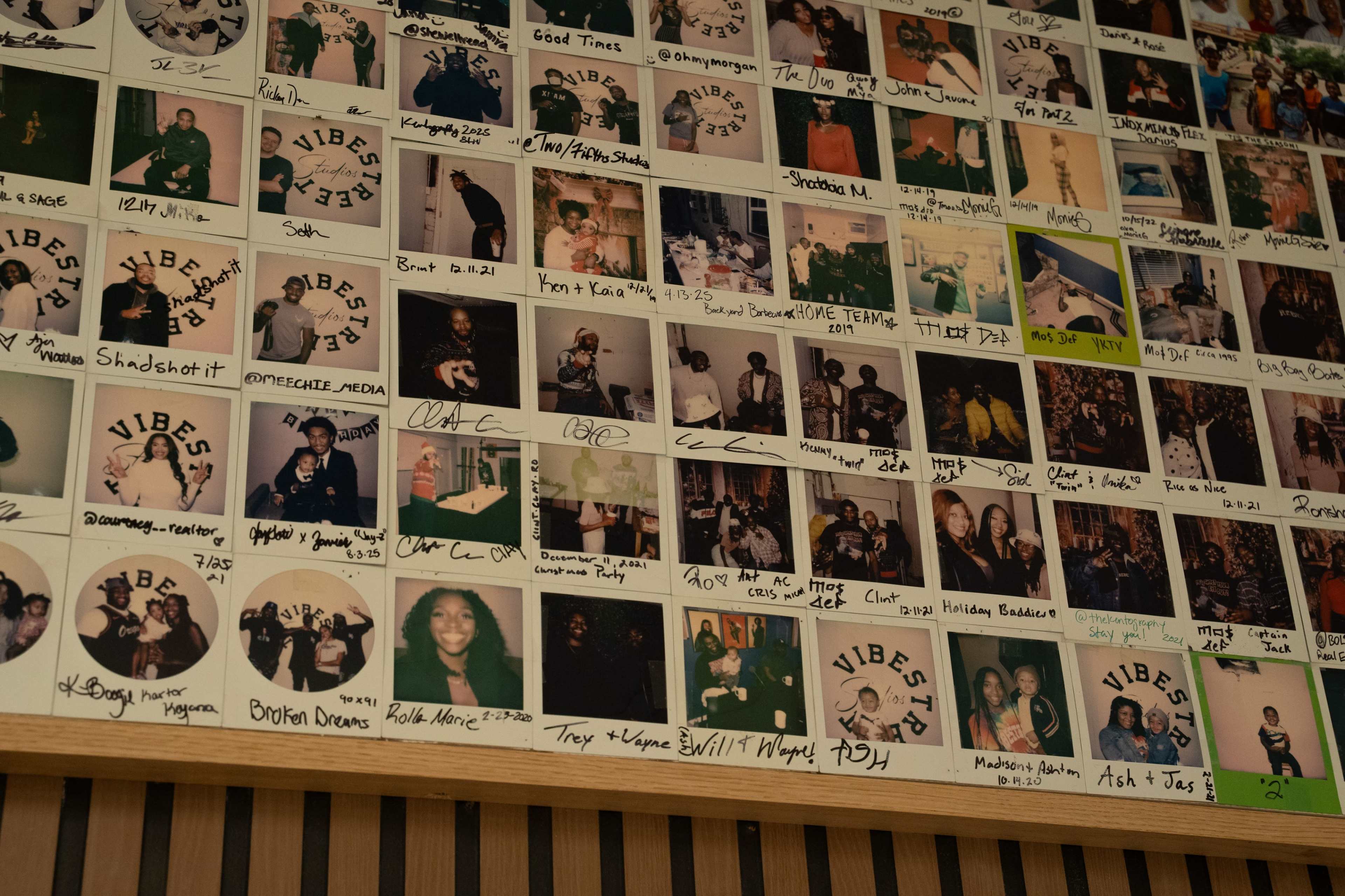The image depicts a wall covered with numerous polaroid-style photographs, each labeled with names and captions, showcasing various people and events.