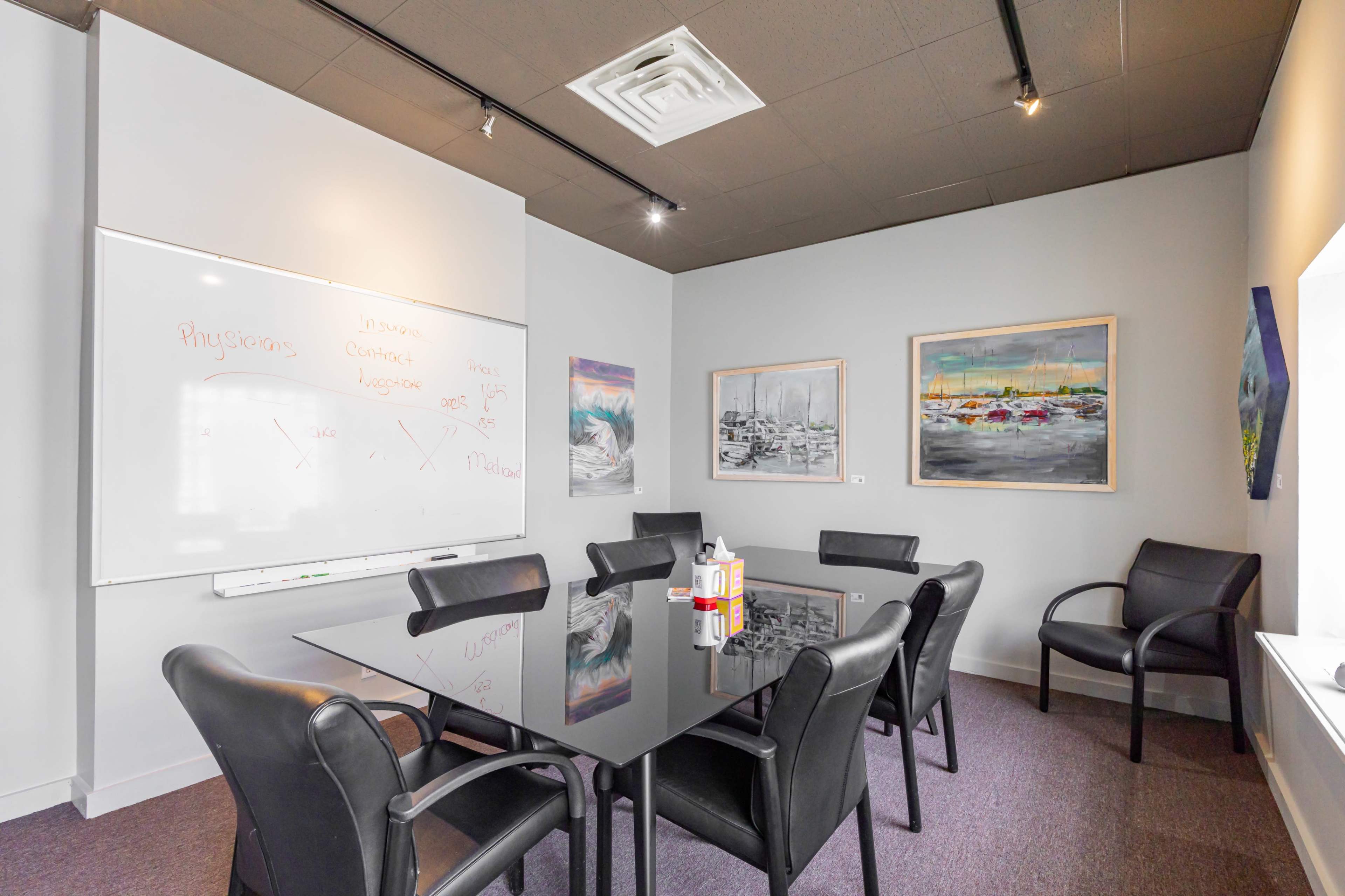 A conference room features a glass table surrounded by black chairs, with a whiteboard on one wall and two paintings hanging on another.