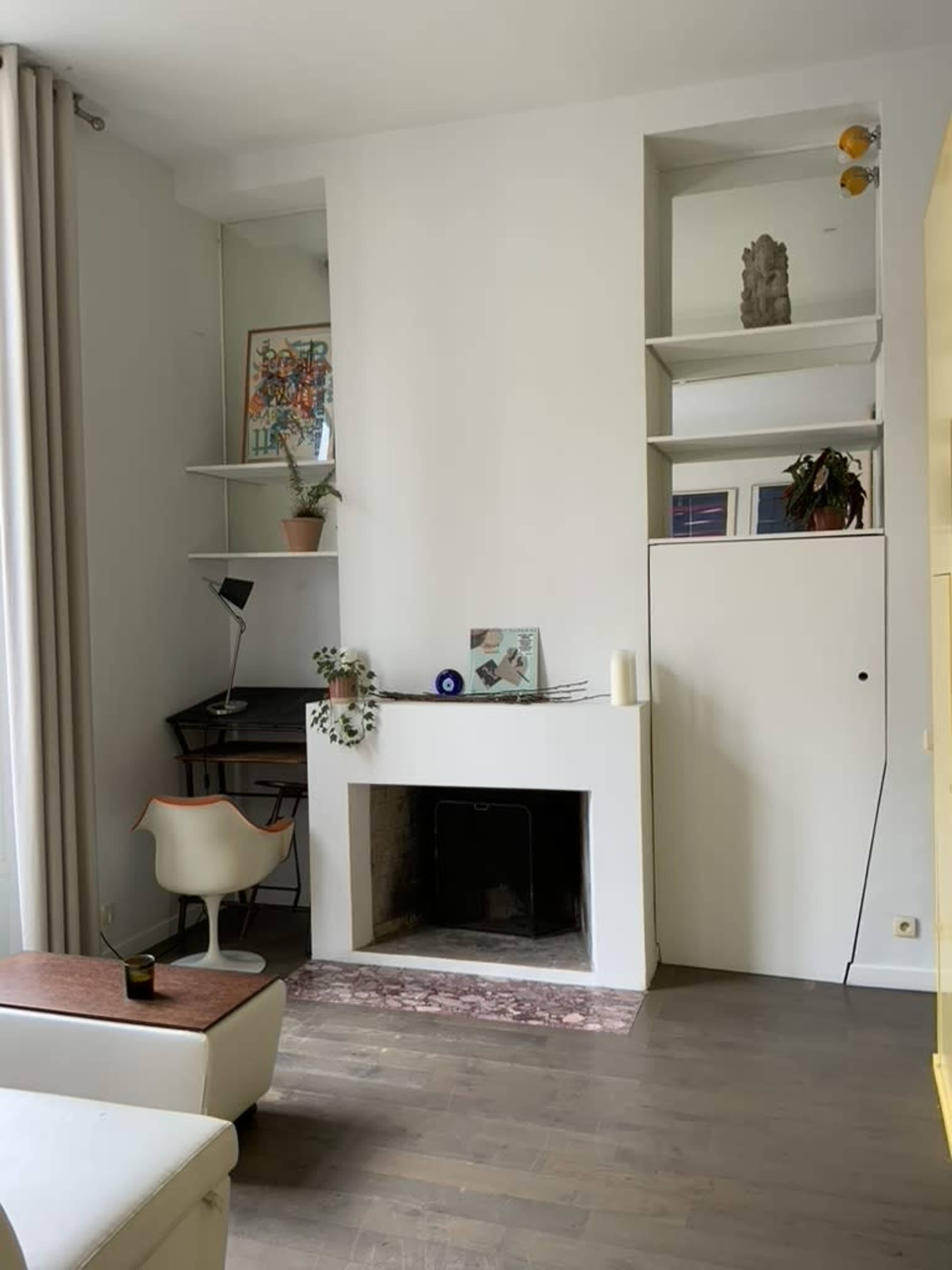 A minimalist living room with a white fireplace, a small desk with a lamp, shelves containing plants and decor, and light wood flooring.