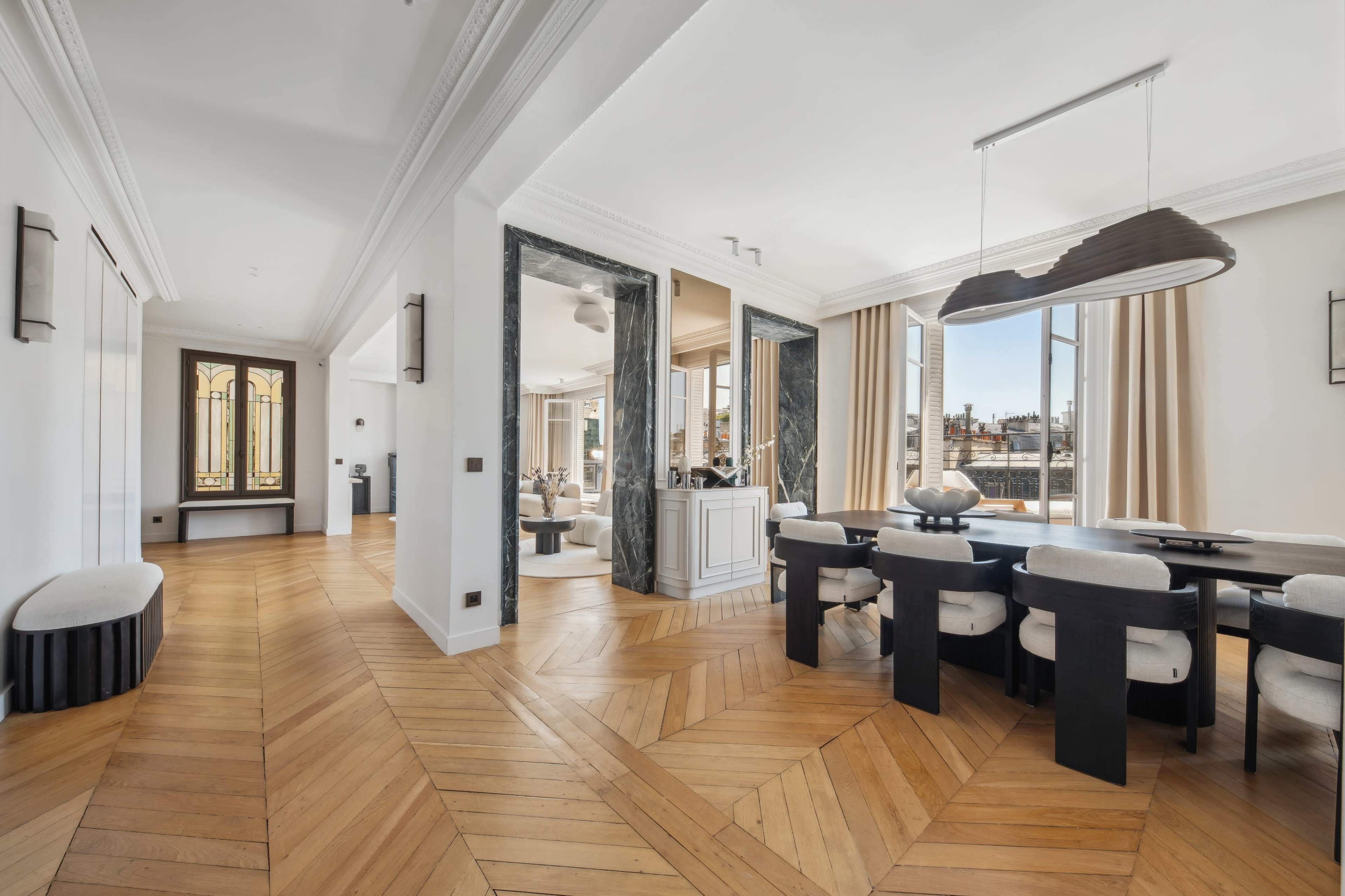 A modern dining area with a large table surrounded by chairs, open to a well-lit living space featuring large windows and a polished wooden floor.