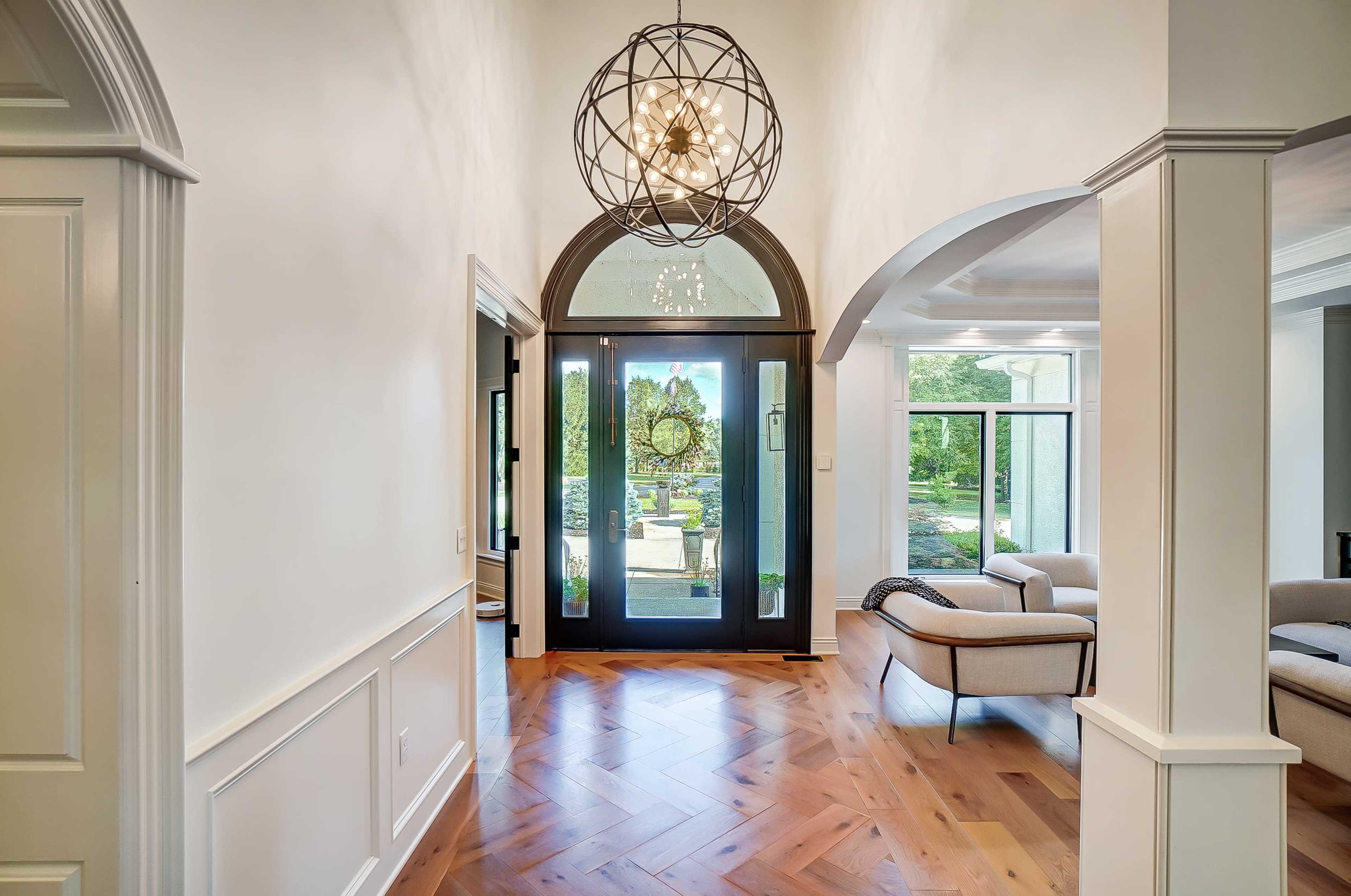 A well-lit foyer with a large arched entryway, featuring a modern chandelier, herringbone-patterned wooden floor, and a view of an outdoor garden through glass doors.