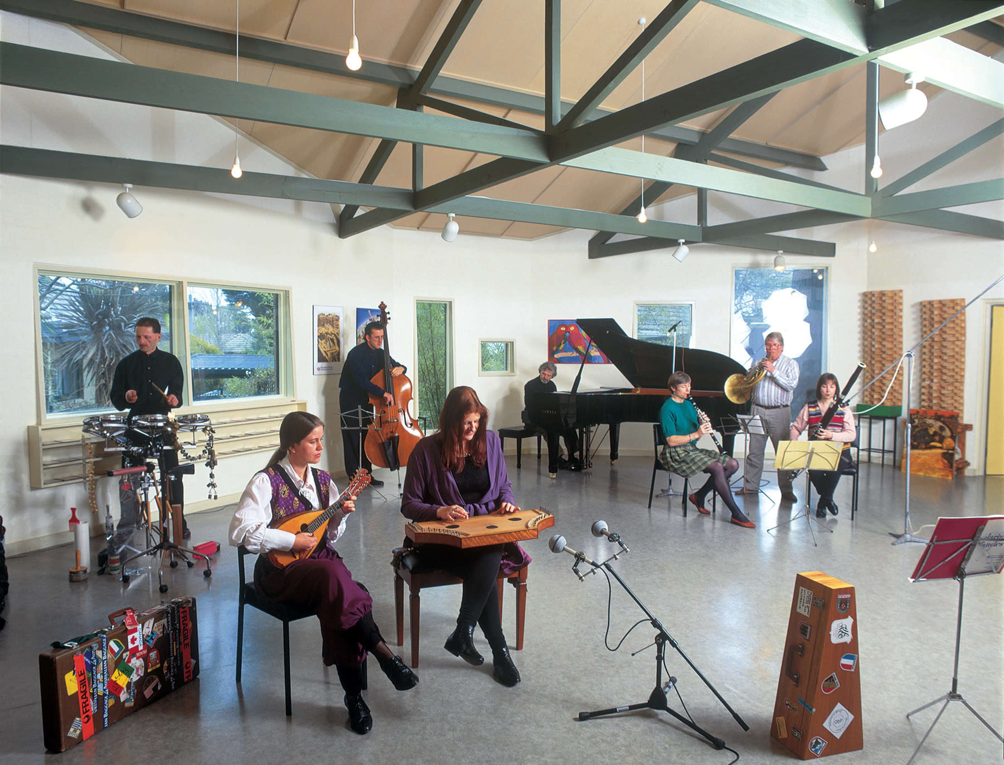 A group of musicians performs together in a brightly lit room with high ceilings and various instruments, including a piano, strings, and brass.