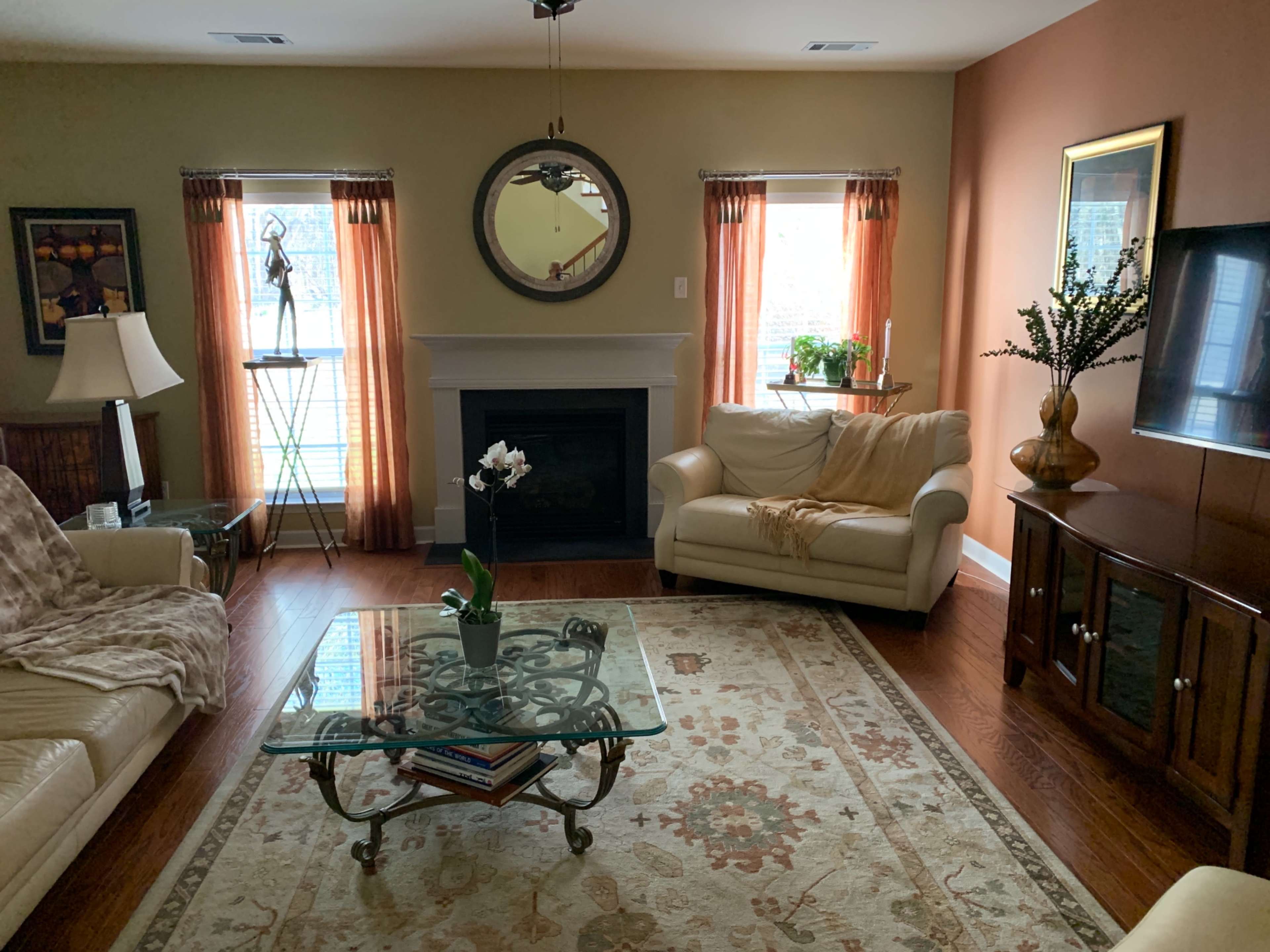 A living room with a beige couch, a glass coffee table on a patterned rug, and windows with orange curtains, all surrounding a fireplace.