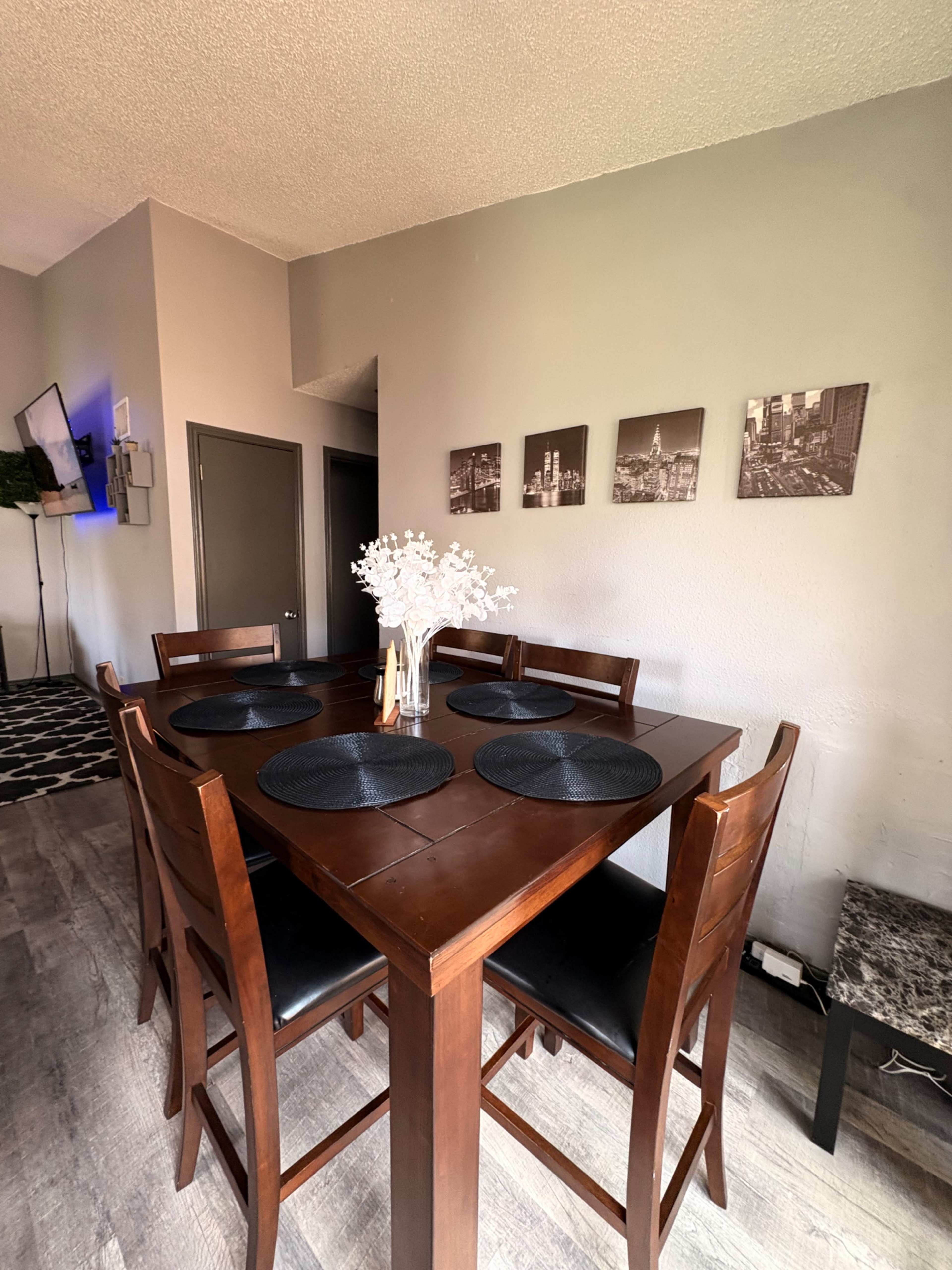 The image shows a wooden dining table with dark placemats and matching chairs, accompanied by a vase of flowers, set against a gray wall adorned with black-and-white photographs.