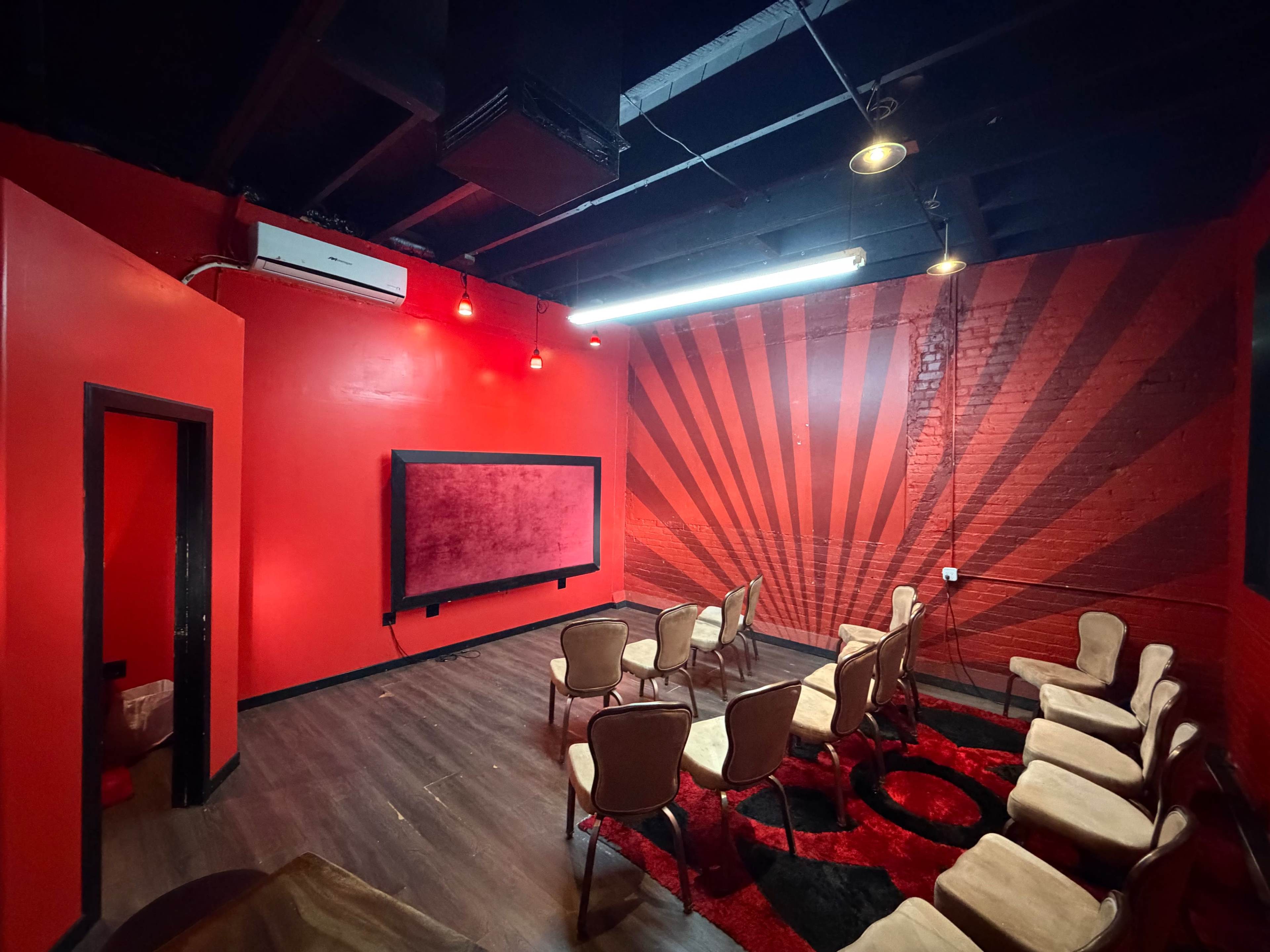 The image shows a small room with red walls featuring a graphic sunburst design, equipped with several chairs arranged in a semi-circle around a large screen.