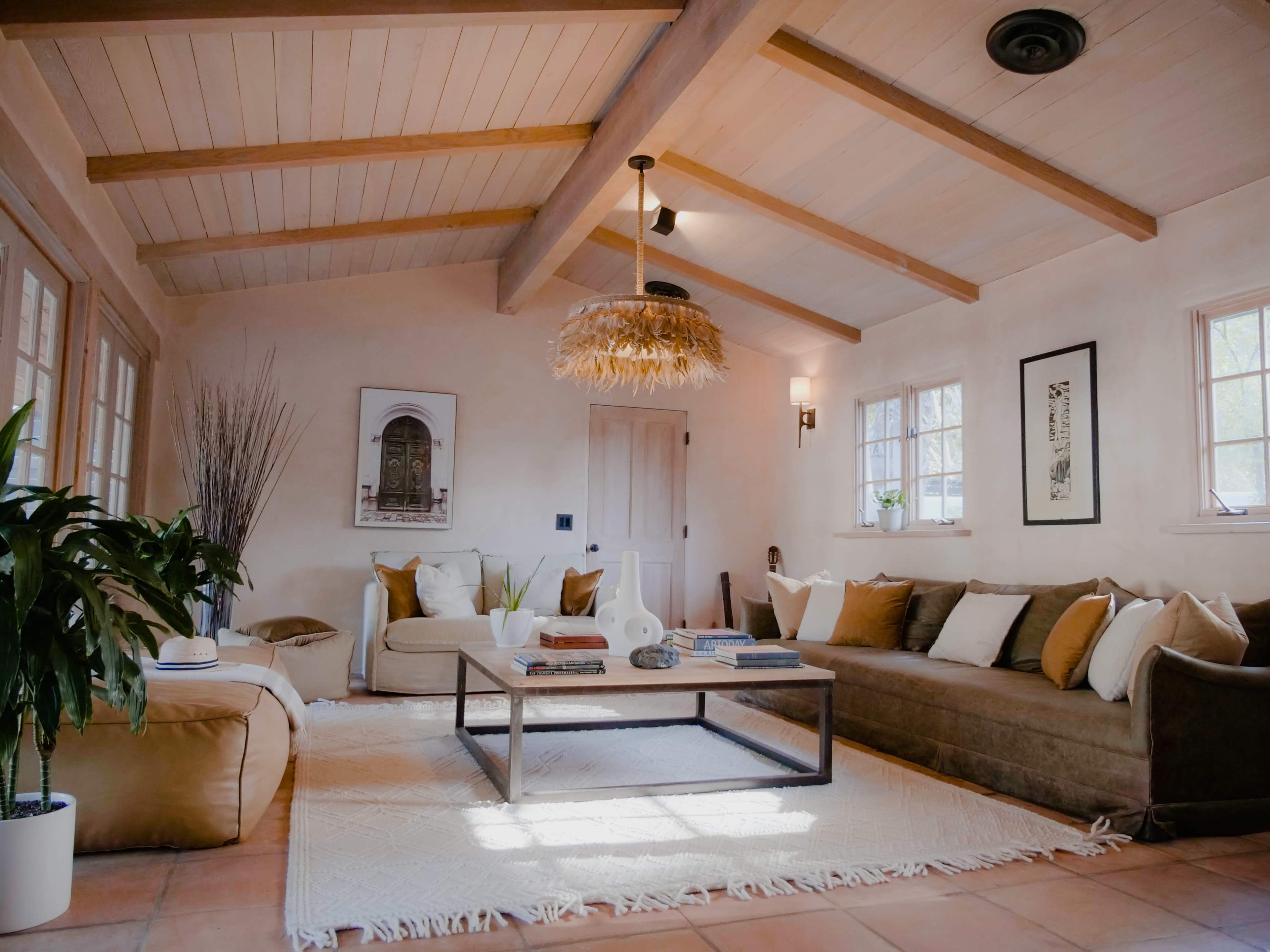 A warmly decorated living room with beige walls, wooden beams, a low coffee table, and a mix of seating arrangements including a sofa and a large floor cushion.