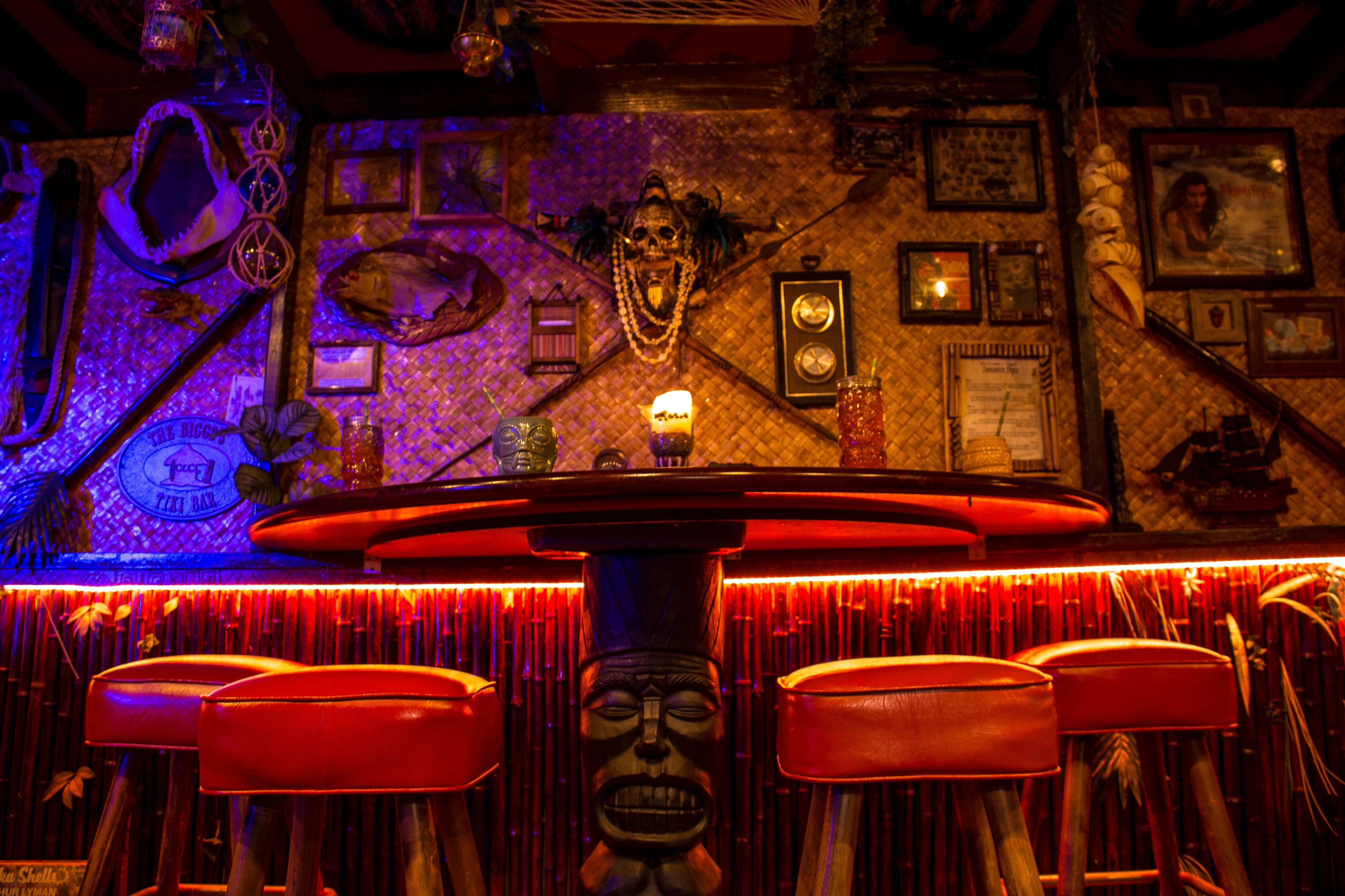 A tiki-themed bar with a round table and four red stools, surrounded by various framed decorations and artifacts on the walls, all illuminated by colorful lighting.
