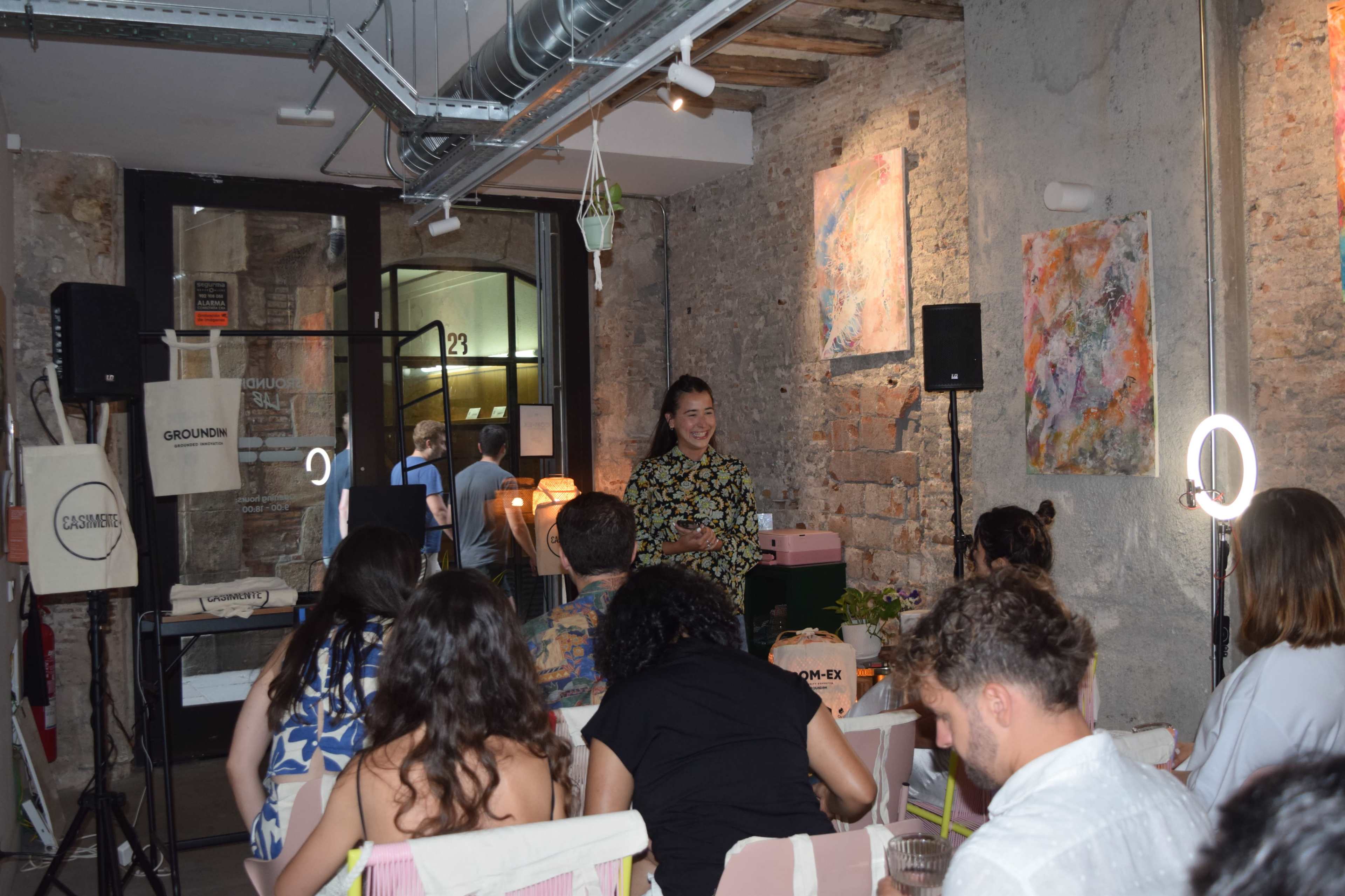 A woman stands in front of an audience in a stylish interior space decorated with artwork, while attendees listen attentively.