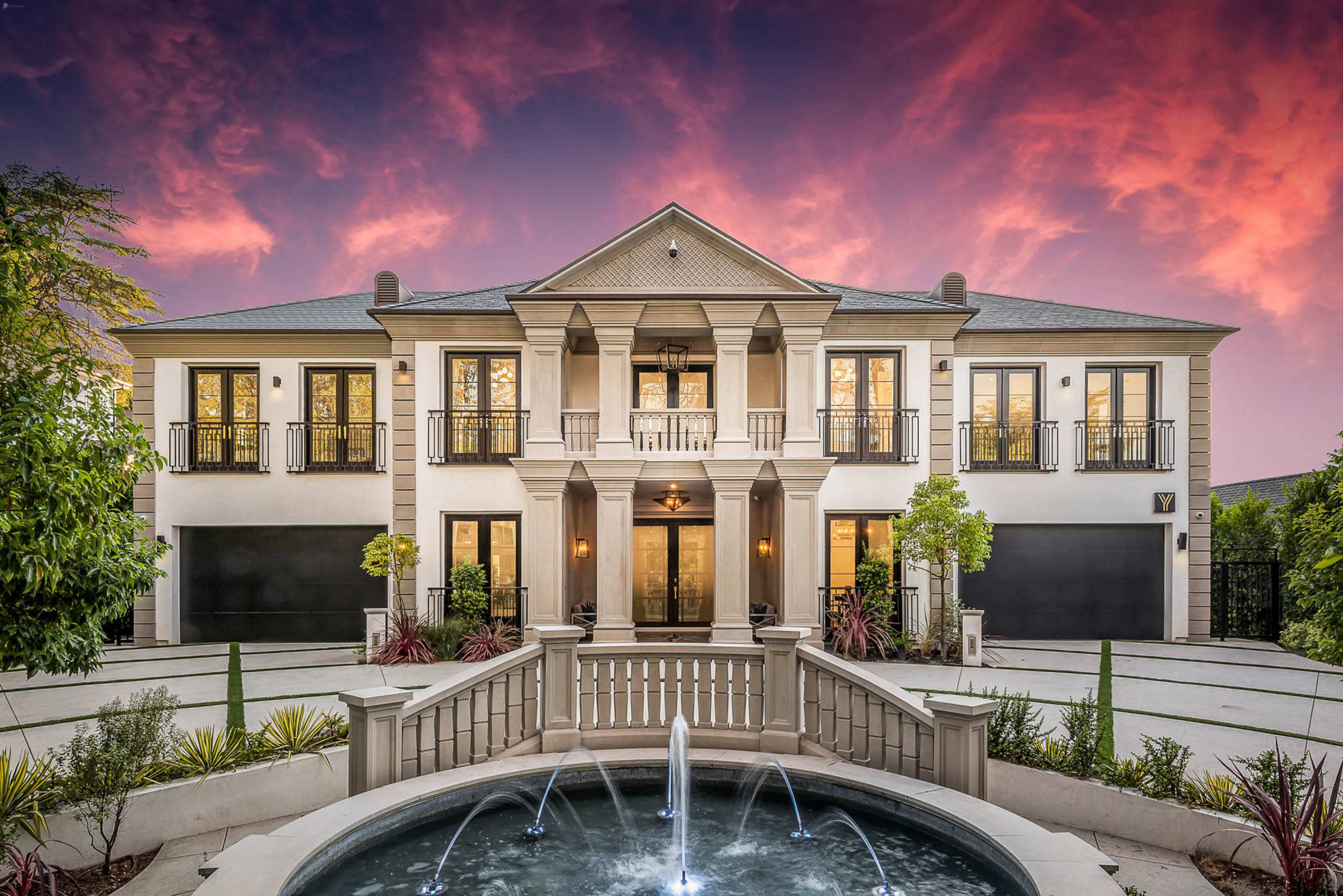 A large, modern mansion with a central fountain, surrounded by manicured landscaping under a vibrant sunset sky.